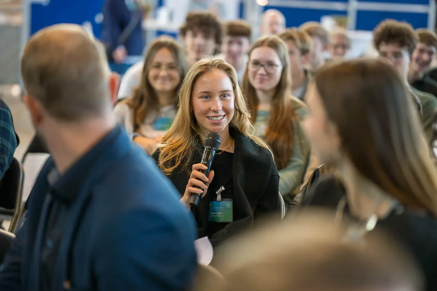 Eine junge Frau mit Mikrofon spricht während einer Veranstaltung vor einem Publikum, das aufmerksam zuhört.