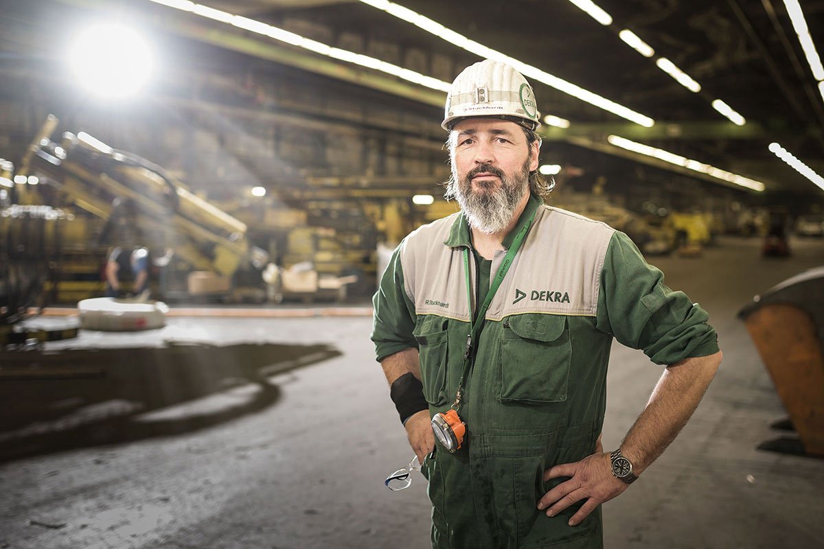 Ein Mann trägt einen Sicherheitshelm und eine Arbeitsweste mit dem Logo DEKRA in einer Fabrikhalle mit schweren Maschinen im Hintergrund.