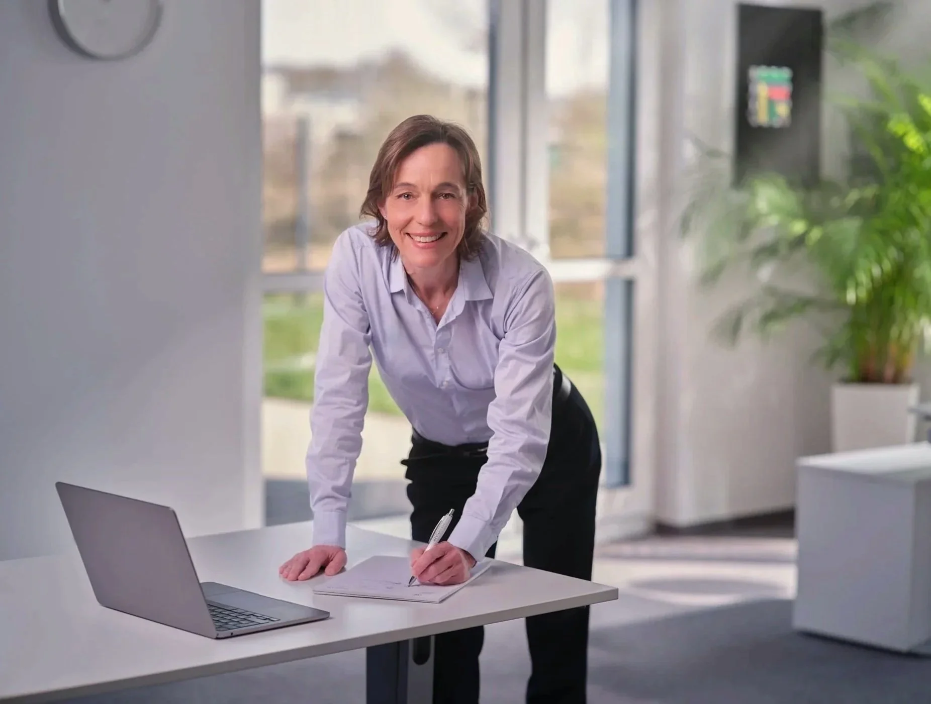 Eine Frau steht an einem weißen Schreibtisch, lächelt in die Kamera und schreibt auf einem Notizblock, während sie ein Laptop vor sich hat. Das Büro ist hell erleuchtet mit großen Fenstern im Hintergrund und Pflanzen als Dekoration.