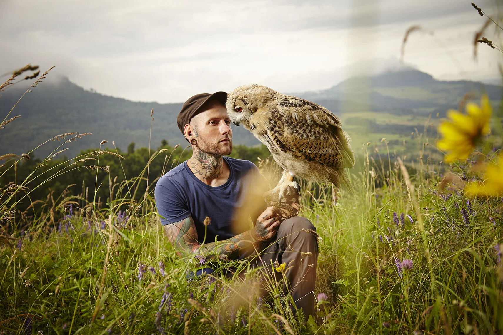 Ein Tattoo-Träger sitzt im Grasfeld, hält eine Eule auf seinem Handschuh und blickt aufmerksam auf die Eule, während die Landschaft im Hintergrund Berge und bewölkten Himmel zeigt.