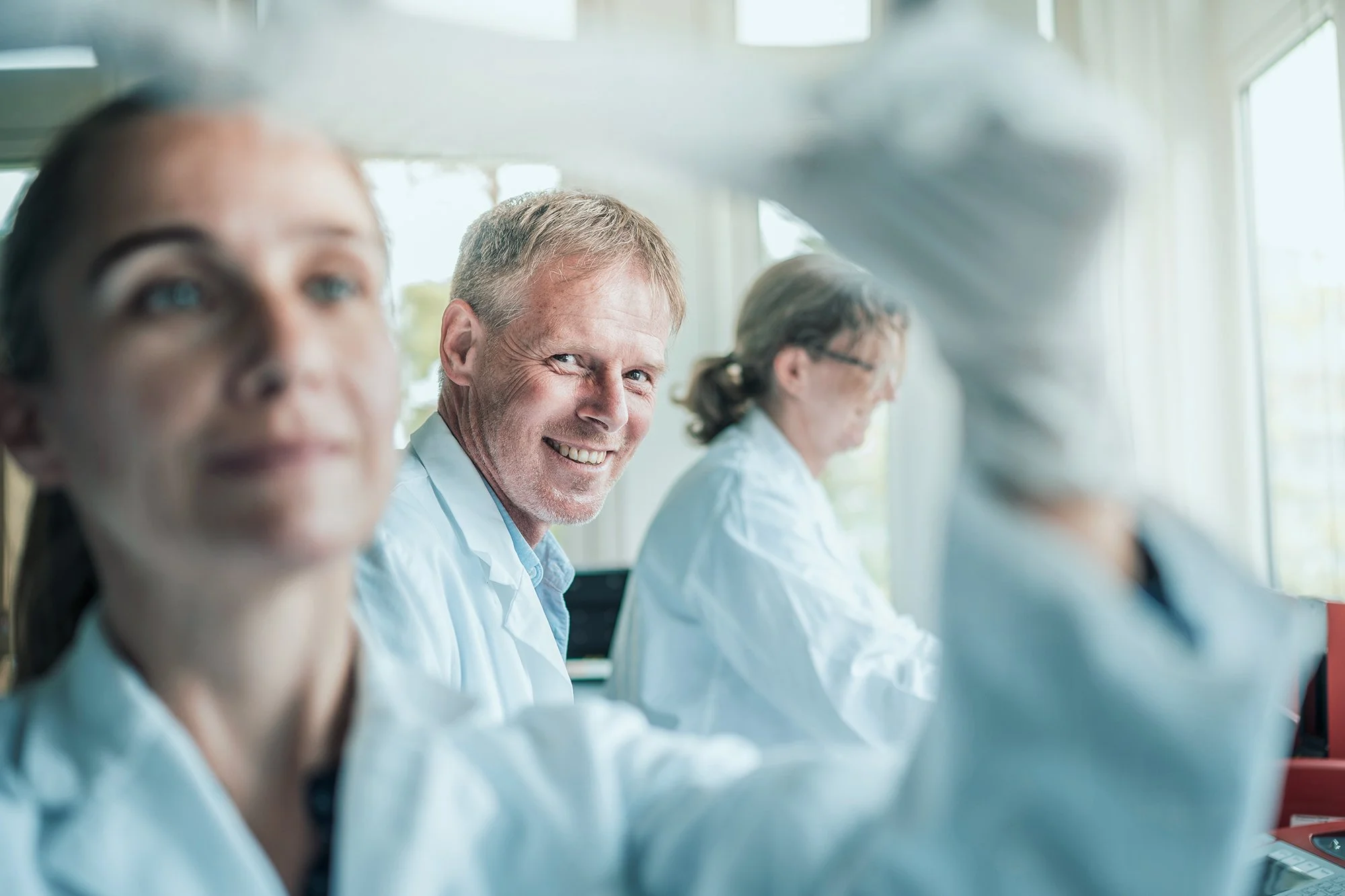 Drei Wissenschaftlerinnen und Wissenschaftler in weißen Laborkitteln sitzen in einem hellen Labor und arbeiten an einem Projekt. Der Mann in der Mitte lächelt freundlich in die Kamera.