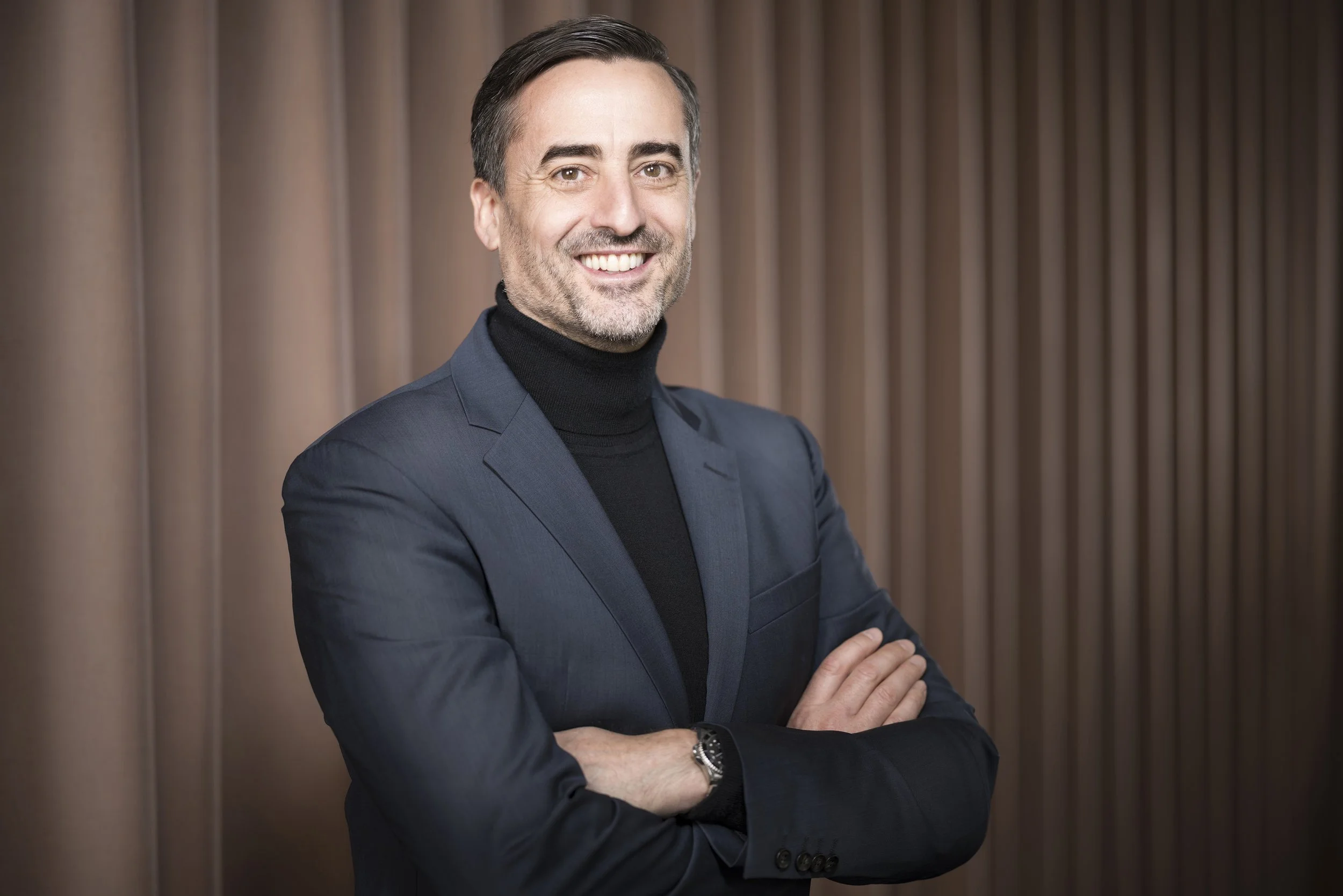 Ein Mann in einem dunklen Anzug und Rollkragenpullover steht vor einem Hintergrund mit vertikalen, braunen Vorhängen, lächelt und hat die Arme verschränkt.