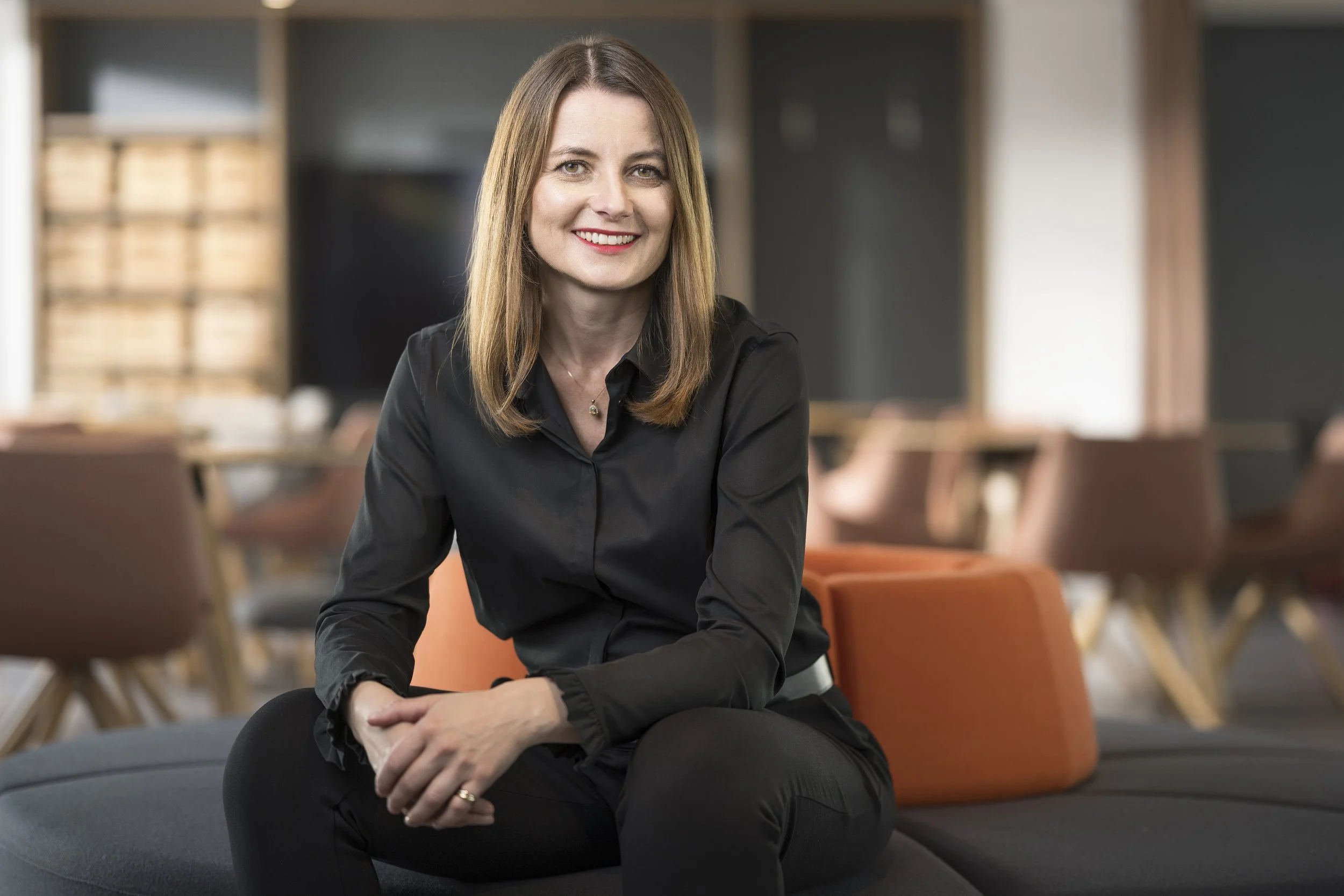 Eine lächelnde Frau mit braunem, geradem Haar, gekleidet in eine schwarze Bluse, sitzt auf einem schwarzen Lounge-Sofa in einem modernen, hellen Raum mit Stühlen im Hintergrund.