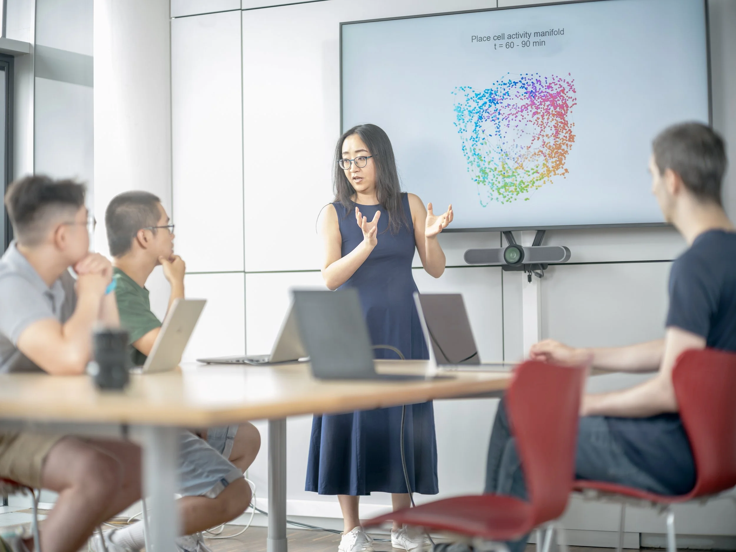 Eine Frau hält eine Präsentation vor einer Gruppe von vier jungen Männern in einem modernen Konferenzraum. Auf einem großen Bildschirm hinter ihr ist ein farbiger, künstlerischer Graph in Herzform mit der Beschriftung 'Place cell activity manifold, t = 60 - 90 min' zu sehen.