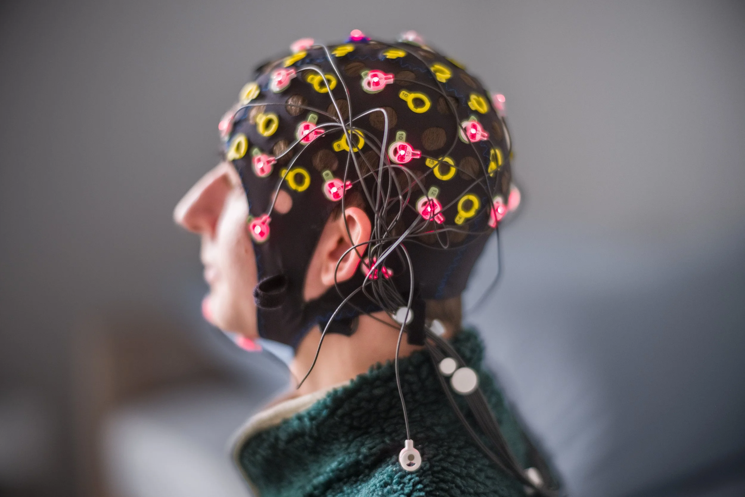 Ein Mensch trägt ein schwarzes EEG-Kappe mit vielen kleinen pinken, gelben und weißen Elektroden, die elektrische Signale messen, während er nach links schaut.