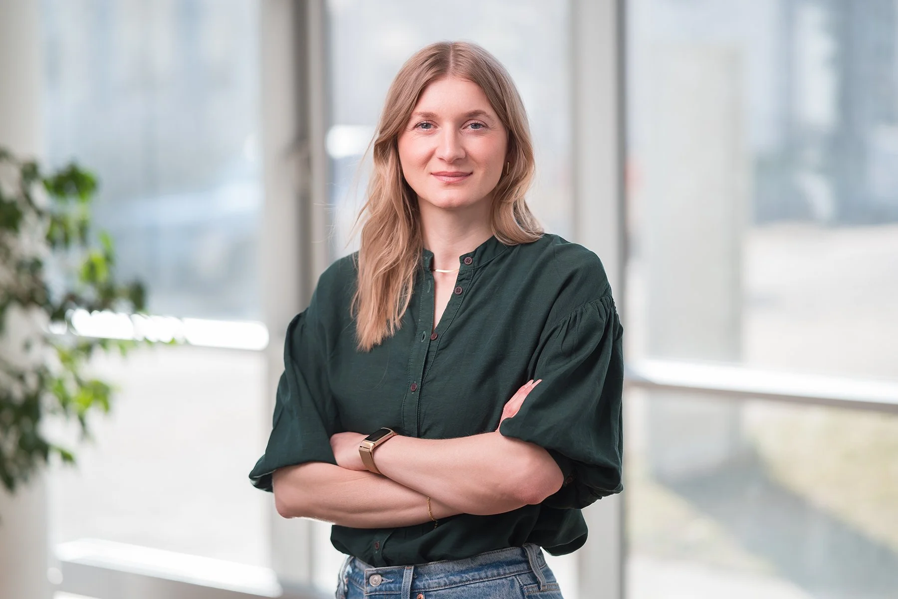 Junge Frau mit crossed arms vor Fenstern in einem Raum, trägt dunkle Bluse, Jeans und eine Smartwatch.