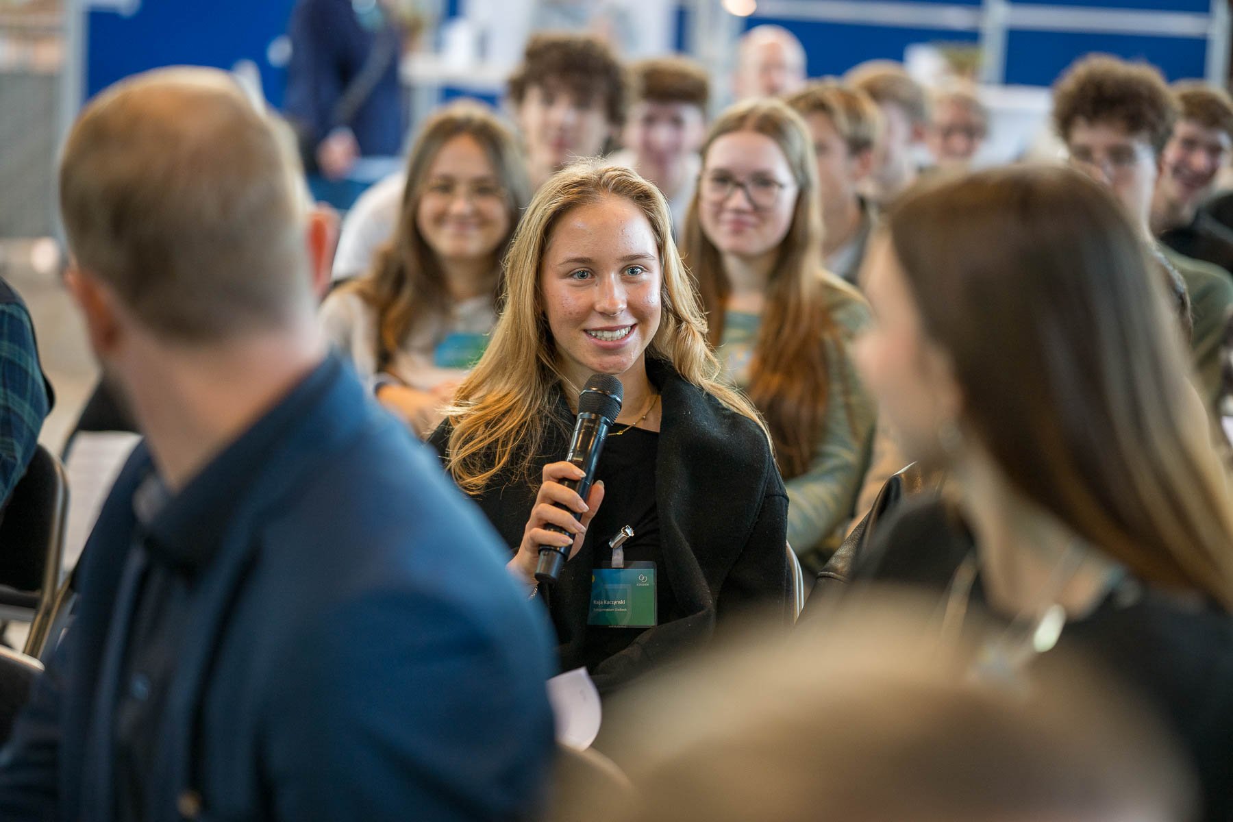 Junge Frau mit Mikrofon in einem Gespräch bei einer Konferenz, im Hintergrund junge Menschen, die zuhören und lachen.
