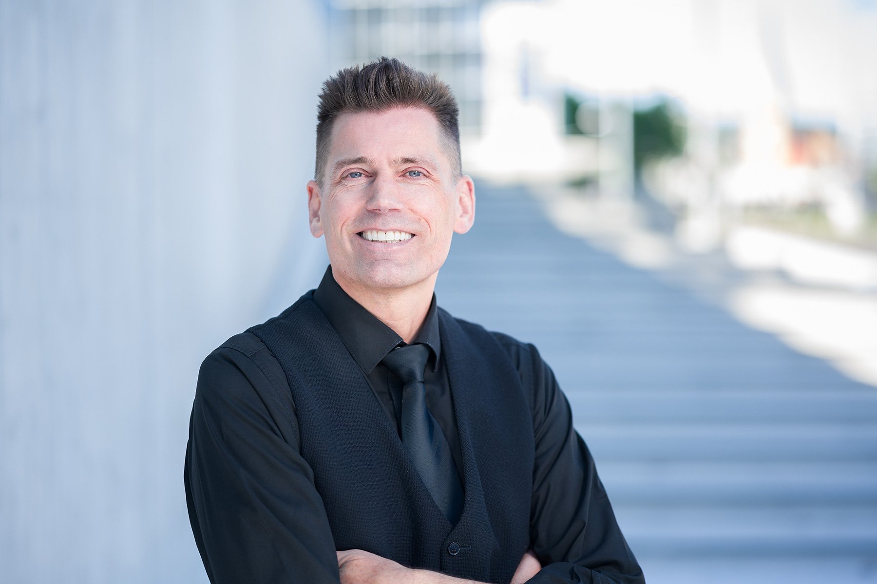 Ein Mann in schwarzem Hemd, Weste und Krawatte steht draußen vor einem modernen Gebäude mit Armlehnen und lächelt in die Kamera.