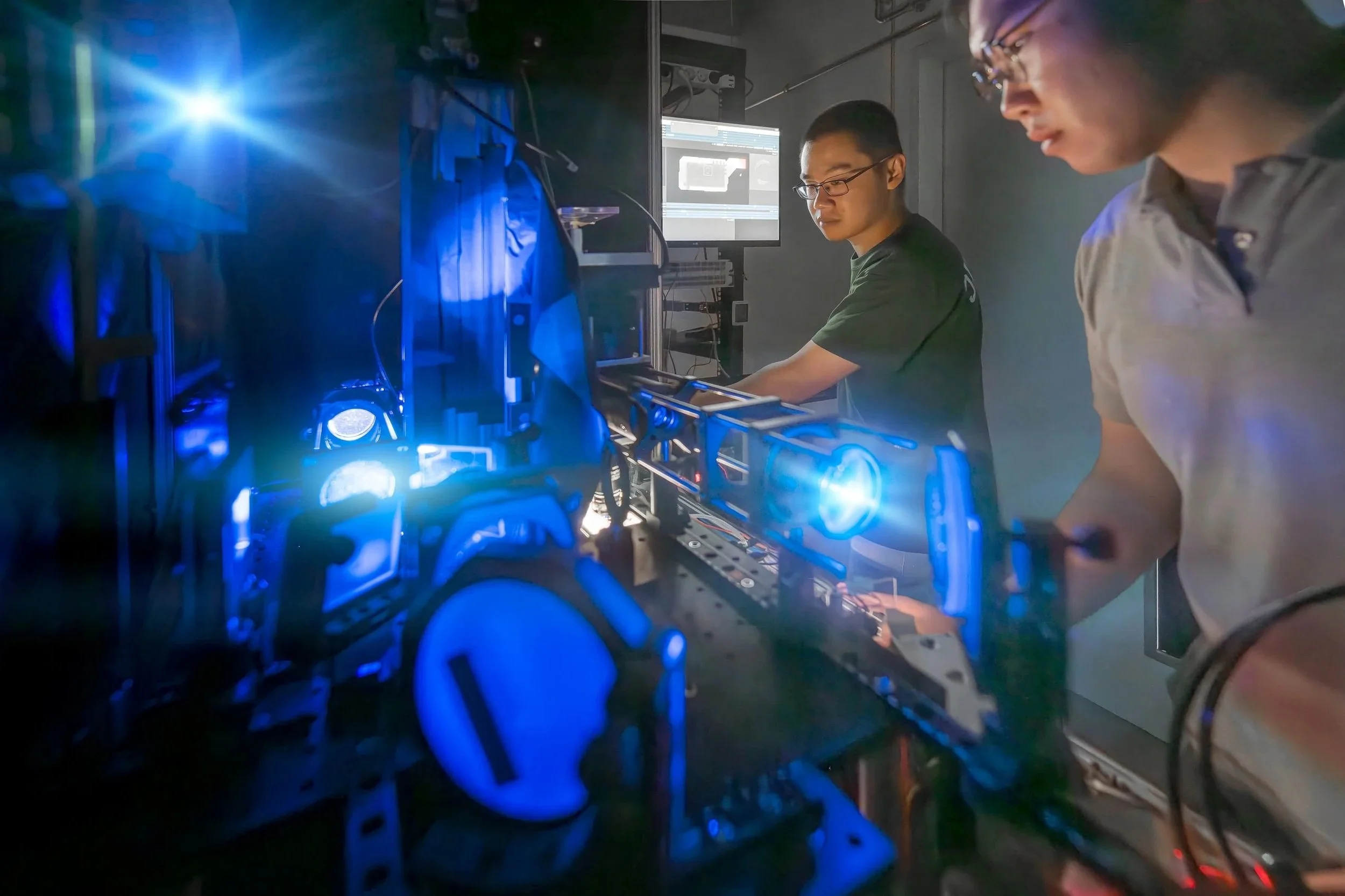Zwei Wissenschaftler arbeiten in einem Labor mit blauem Licht an einem technischen Gerät, während im Hintergrund ein Computerbildschirm zu sehen ist.