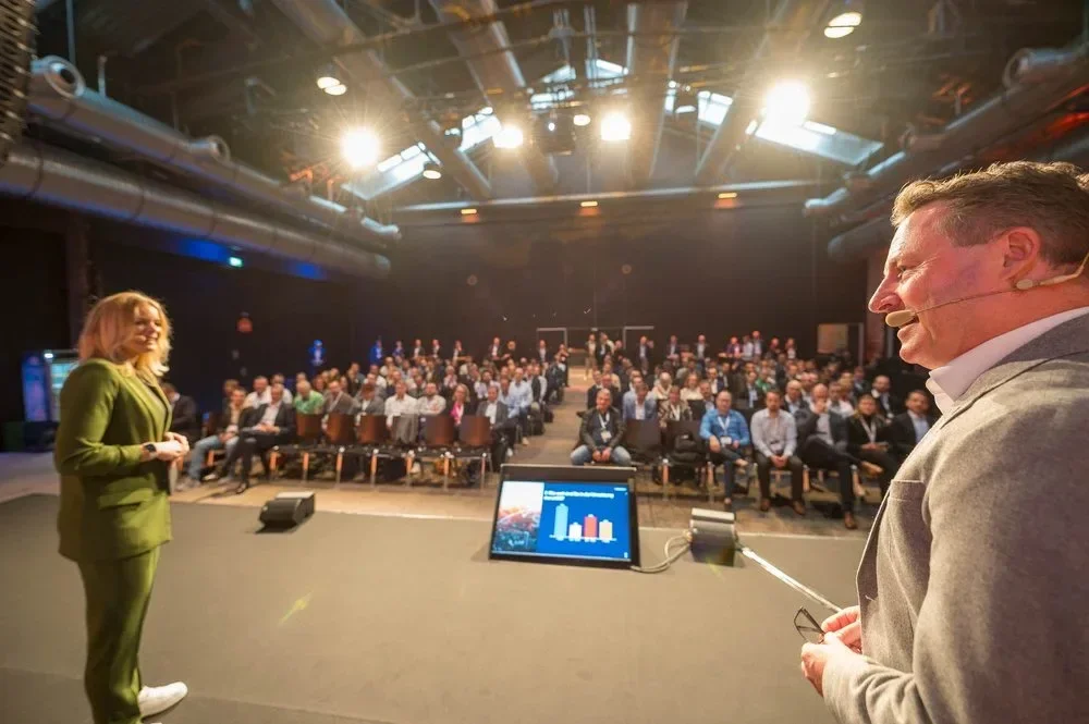 eine Frau hält eine Präsentation vor einem Publikum in einem großen Saal mit professionellem Bühnenlicht, während ein Moderator mit Mikrofon zuhört.