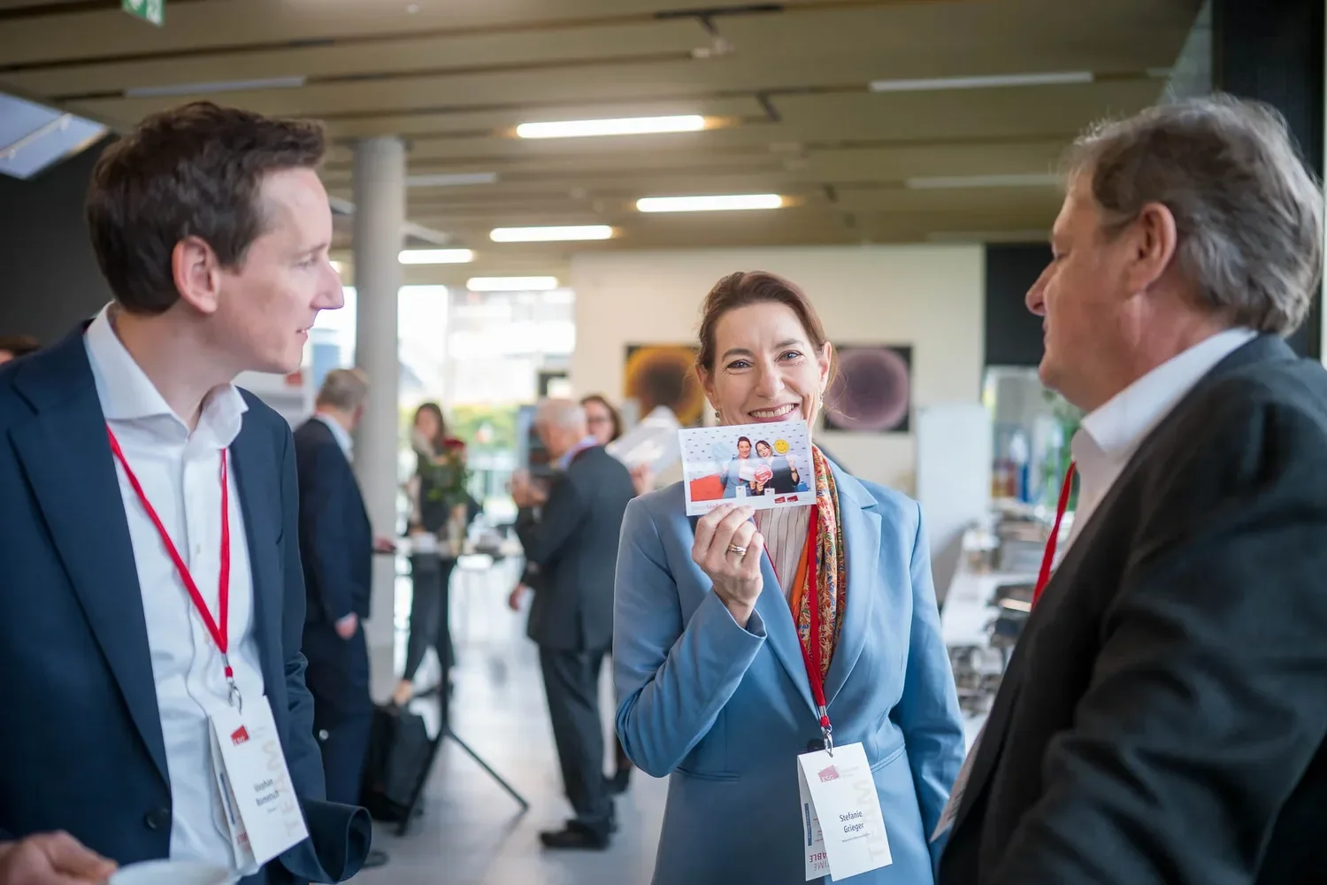 Drei Personen im Gespräch bei einem Geschäftstreffen, eine Frau hält eine ID-Karte und lächelt.