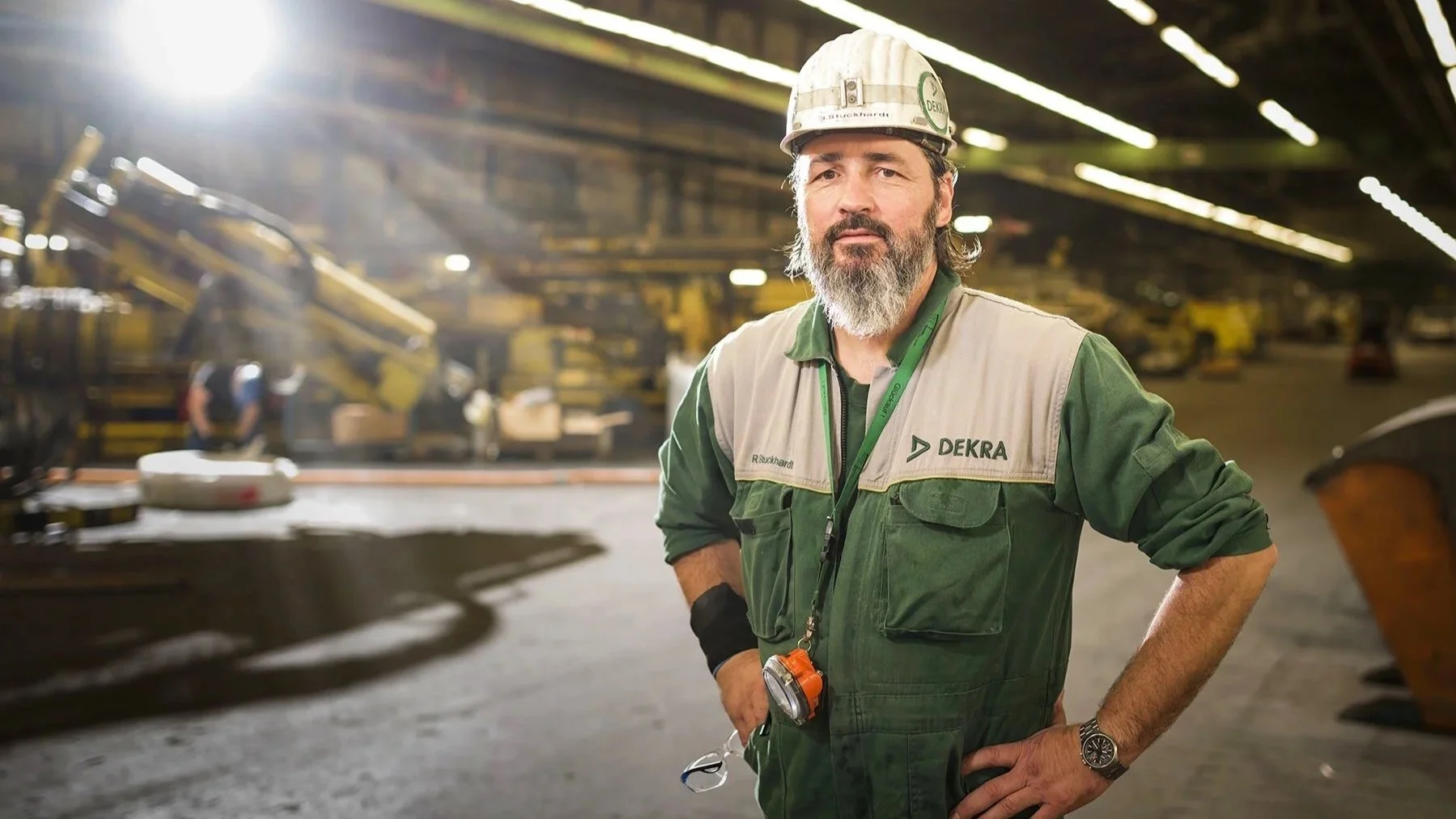 Ein Mann in Sicherheitskleidung und Helm steht in einer Fabrikhalle mit Maschinen im Hintergrund.