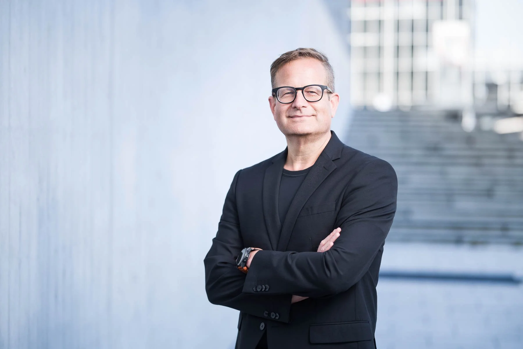 Ein Mann in dunklem Anzug und Brille steht vor einer modernen, grauen Wand und lächelt leicht mit verschränkten Armen.