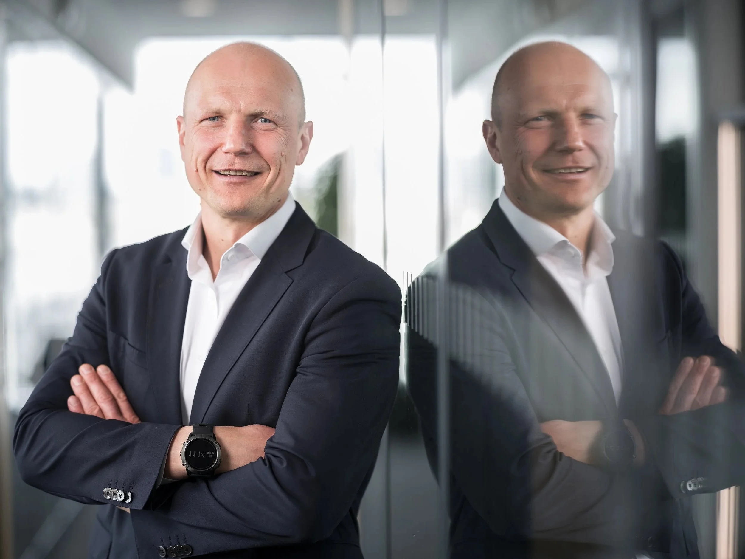 Ein Mann in einem schwarzen Anzug und weißem Hemd steht vor einer Glaswand, lächelt und hat die Arme verschränkt.