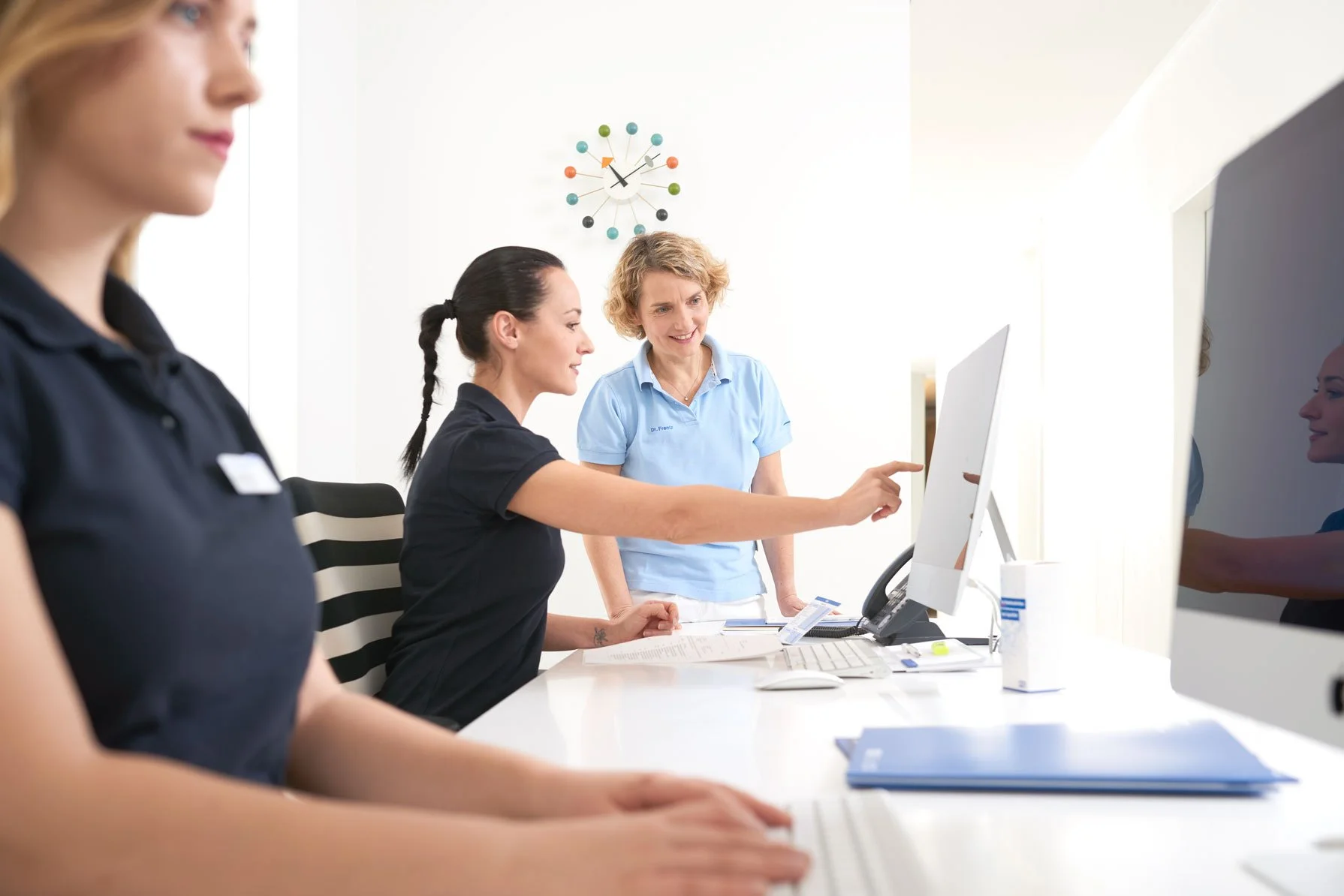 Drei Frauen in einem Büro, eine sitzt am Computer, eine zeigt auf den Bildschirm, eine steht daneben und erklärt etwas.