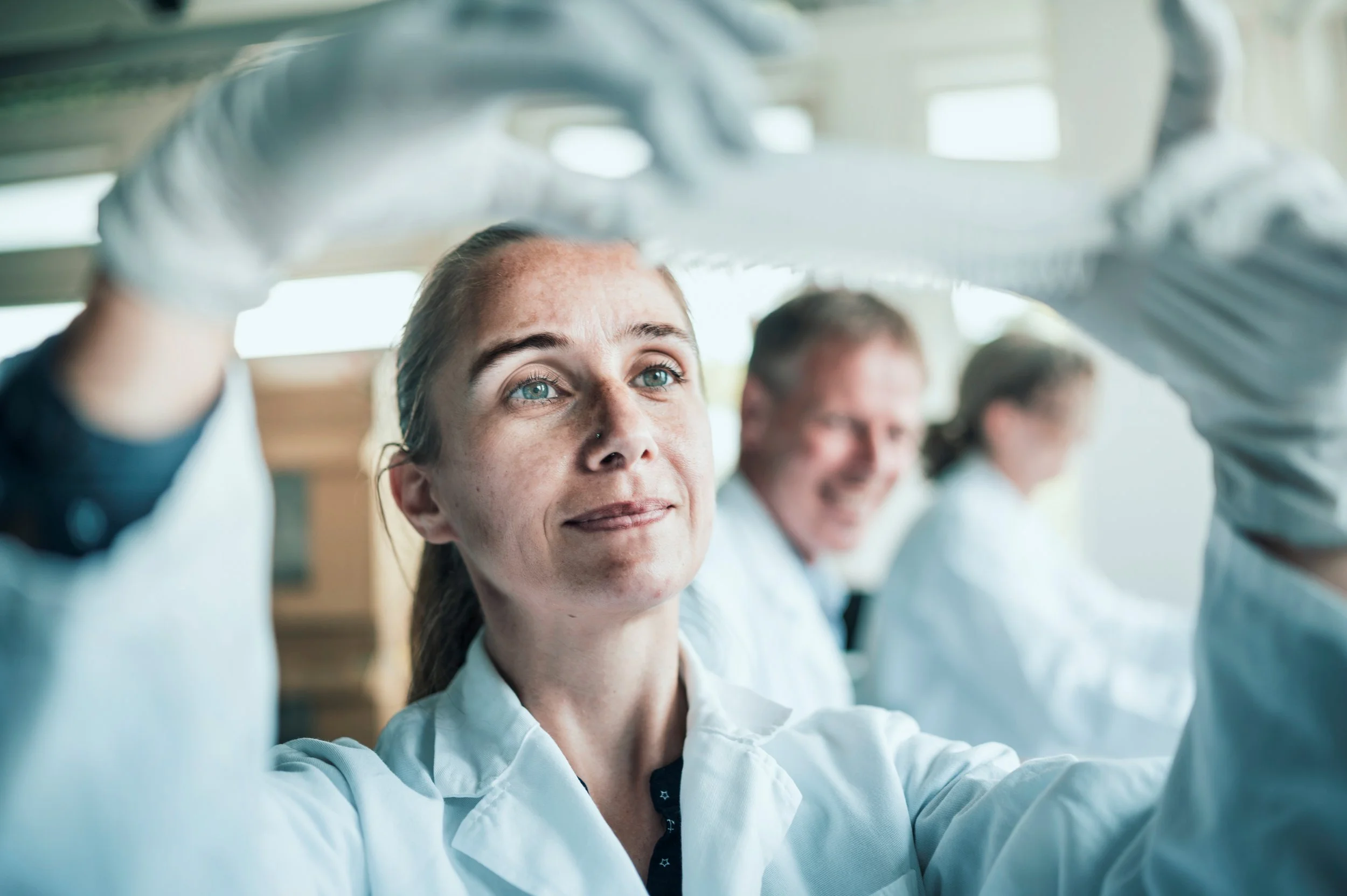 Frau trägt eine Schutzbrille und eine weiße Jacke, arbeitet in einem Labor, während andere Personen im Hintergrund auch arbeiten.