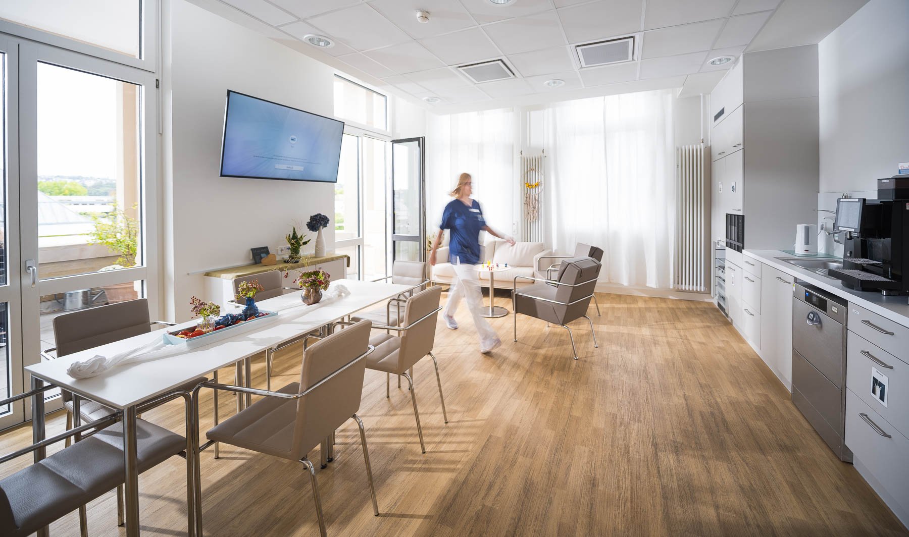 Ein modern eingerichteter Gemeinschaftsraum oder Pausenraum mit einem langen weißen Tisch, mehreren Stühlen, einem Sofa, einem Bildschirm an der Wand, einer Person in blauer Uniform, die sich im Raum bewegt, und Küchenzeile mit Kaffeemaschine und Wasserkocher.