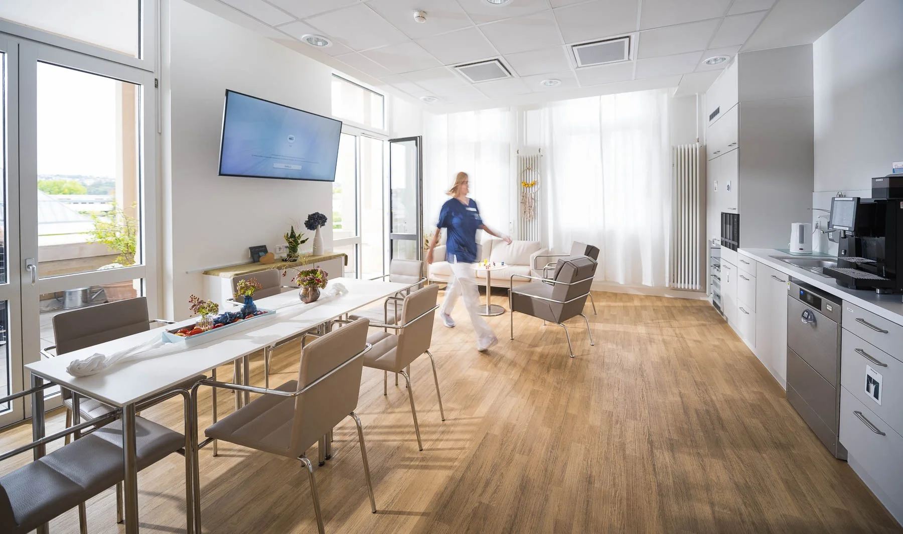 Ein modern eingerichteter Gemeinschaftsraum oder Pausenraum mit einem langen weißen Tisch, mehreren Stühlen, einem Sofa, einem Bildschirm an der Wand, einer Person in blauer Uniform, die sich im Raum bewegt, und Küchenzeile mit Kaffeemaschine und Wasserkocher.