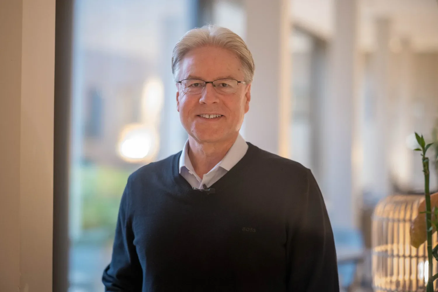Ein älterer Mann mit Brille,lockigem grauem Haar trägt einen weißen Hemd und einen schwarzen Pullover und lächelt vor einem Fenster in einem Raum mit Pflanzen und natürlichem Licht.