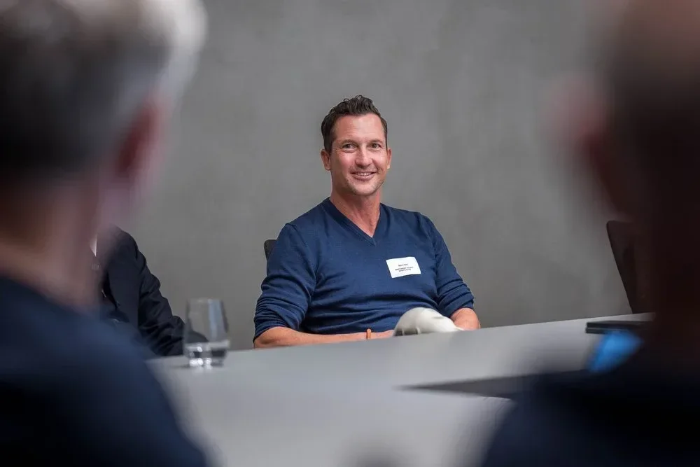 Mann lächelnd bei einer Besprechung, Carl in einem blauen Pullover, im Konferenzraum.