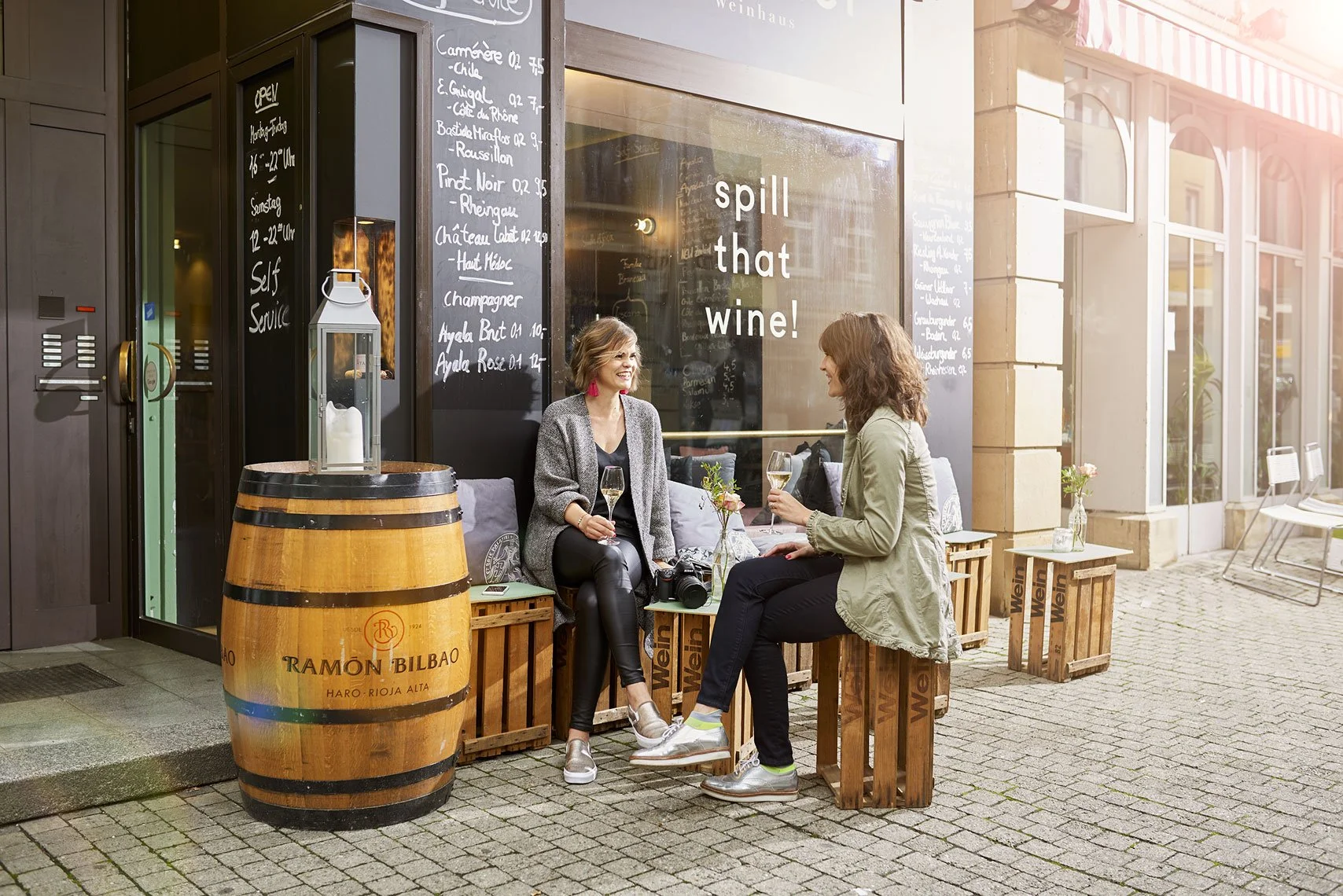 Zwei Frauen sitzen vor einer Weinbar auf Holzkisten und trinken Wein, während sie sich unterhalten. Es ist Nachmittag und die Bar ist im Außenbereich. Es gibt ein Schild mit der Aufschrift »spill that wine!« und eine Weinkarte vor der Bar ist sichtba