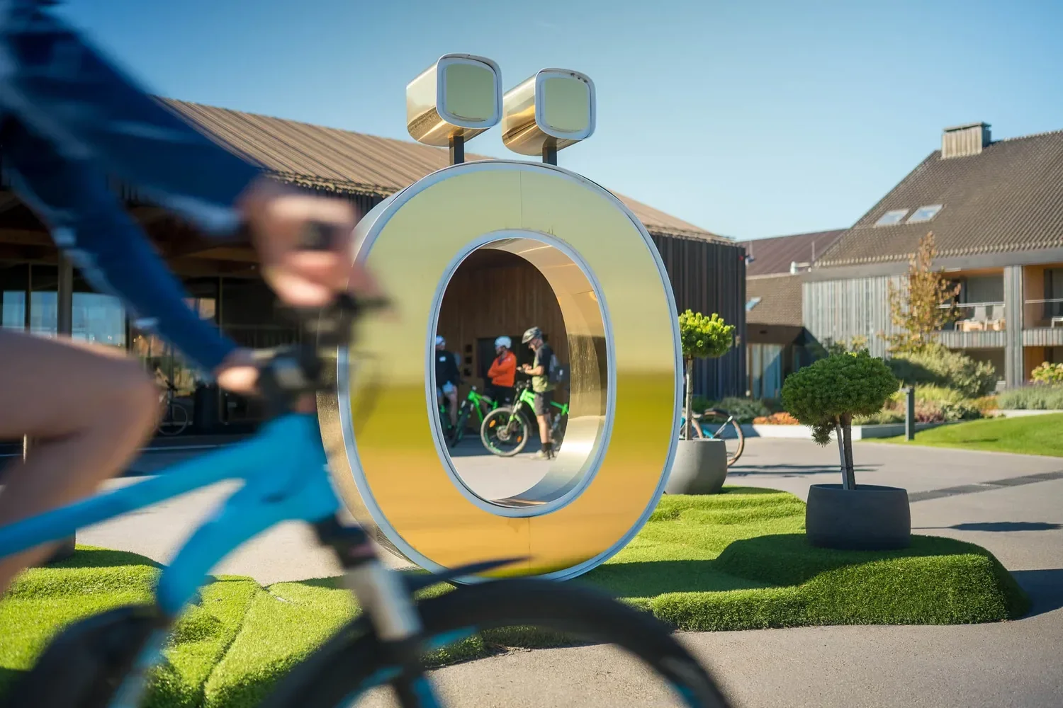 Ein Fahrrad im Vordergrund, reflektiert in einer großen goldfarbenen, ringförmigen Skulptur mit Verbraucher-Design, im Hintergrund sind Menschen mit Fahrrädern und Helmen zu sehen, die auf einem Platz vor modernen Gebäuden stehen.