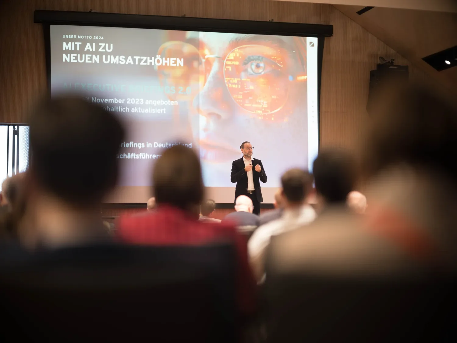Ein Mann hält eine Präsentation vor einem Publikum in einem Saal mit einem großen Bildschirm im Hintergrund, der Text und ein Bild eines Gesichts mit digitaler Augenansicht zeigt.