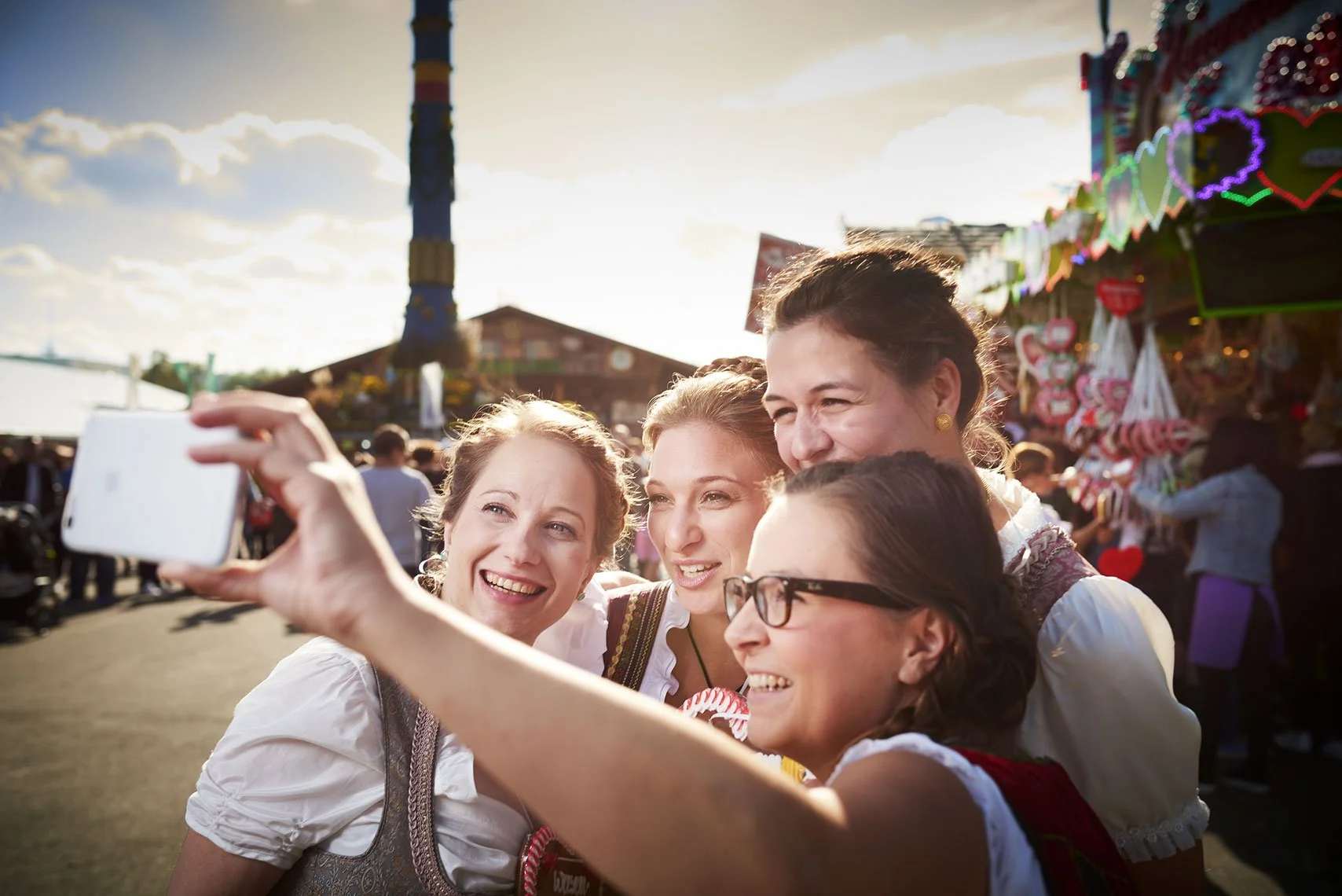 Vier Frauen in traditionellen Trachten machen ein Selfie auf einem Volksfest, im Hintergrund sind Verkaufsstände mit Süßigkeiten und Dekorationen zu sehen, bei Sonnenuntergang.