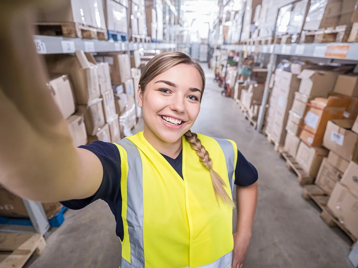 Eine lächelnde junge Frau in einer gelben Sicherheitsweste macht ein Selfie in einem Lager mit Regalen voller Pakete.