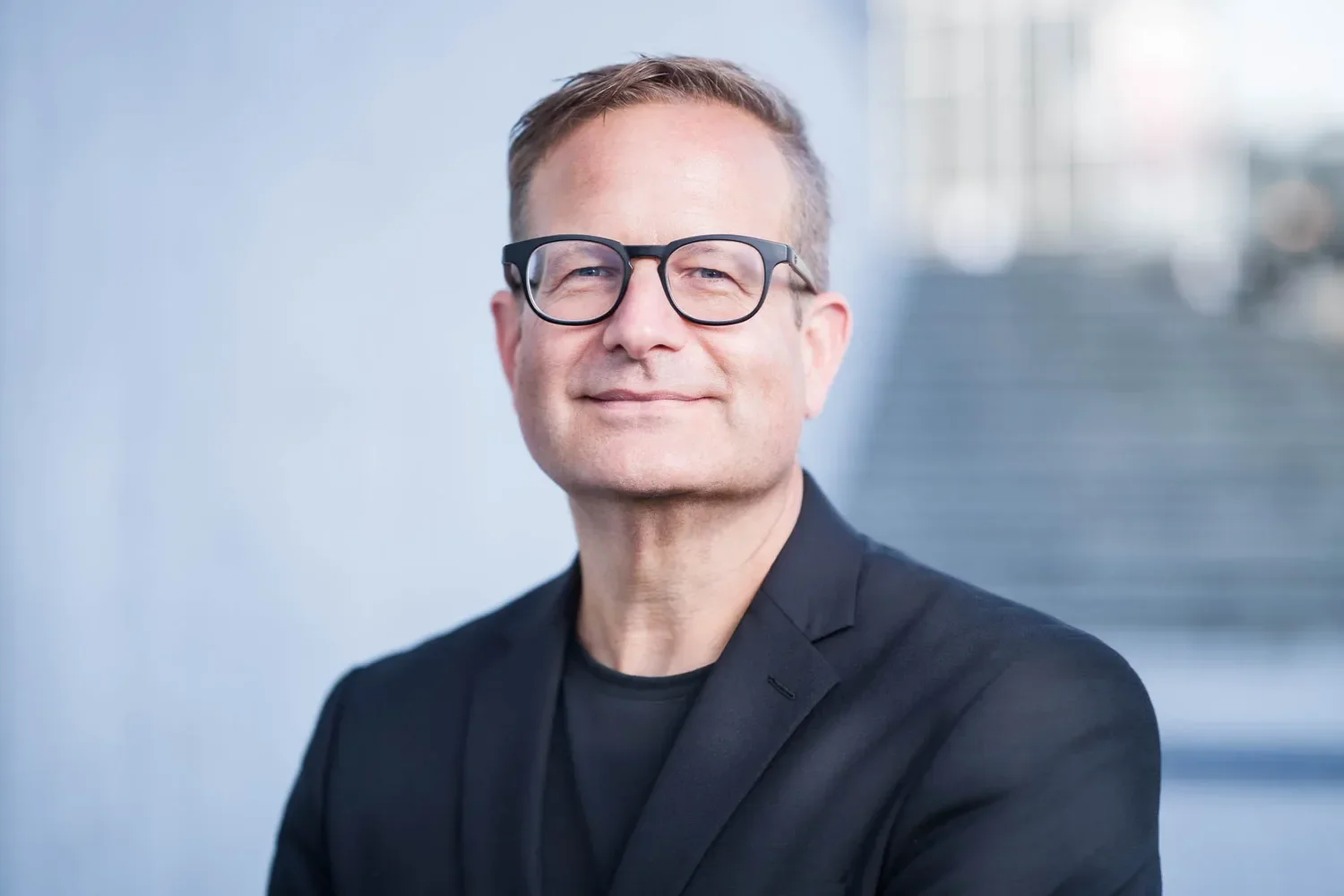 Ein Mann mit Brille trägt einen schwarzen Anzug und lächelt. Er steht vor einem Hintergrund mit einem modernen Gebäude.