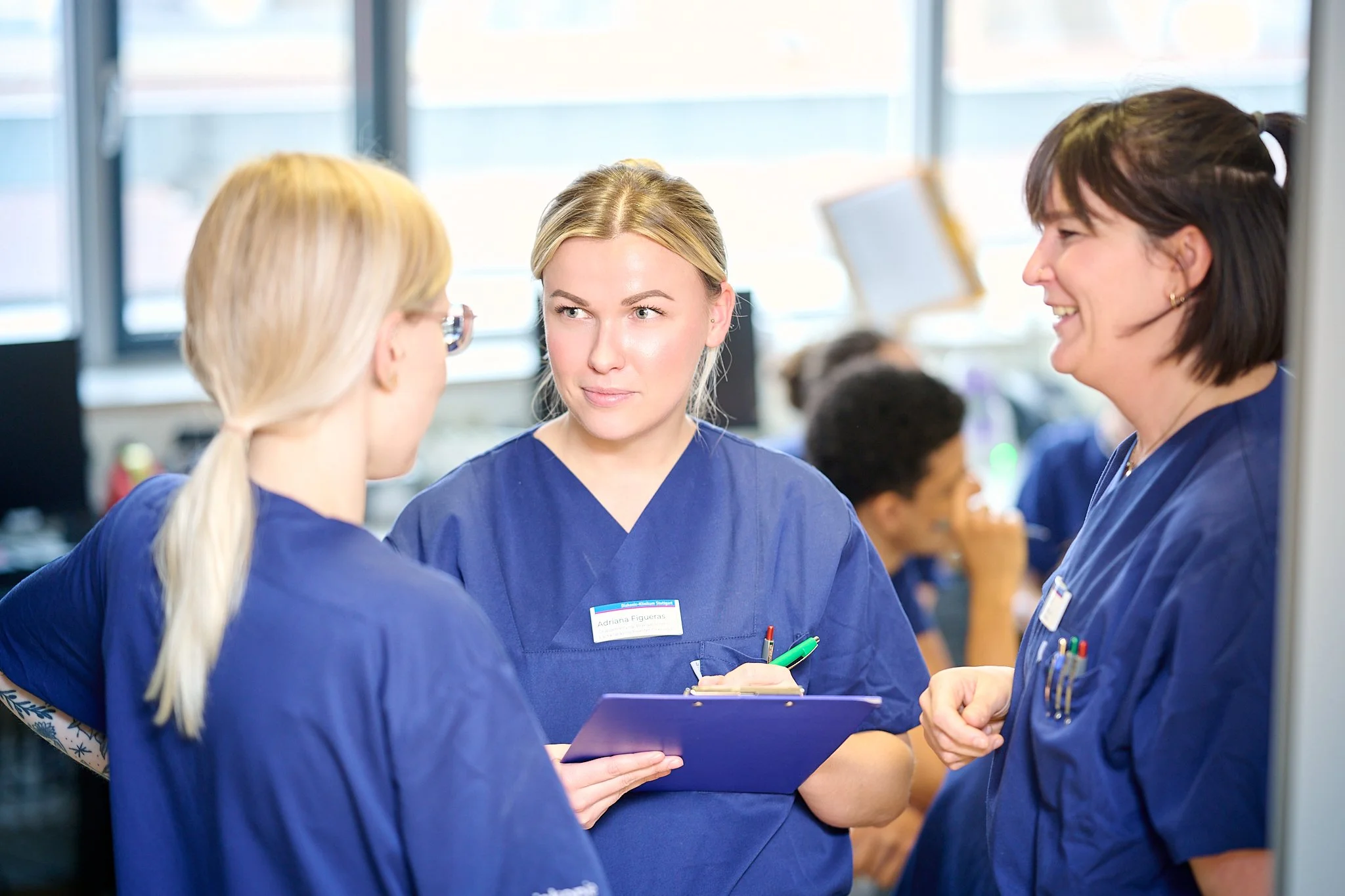 Drei Krankenschwestern in blauer Uniform unterhalten sich in einem Krankenzimmer.