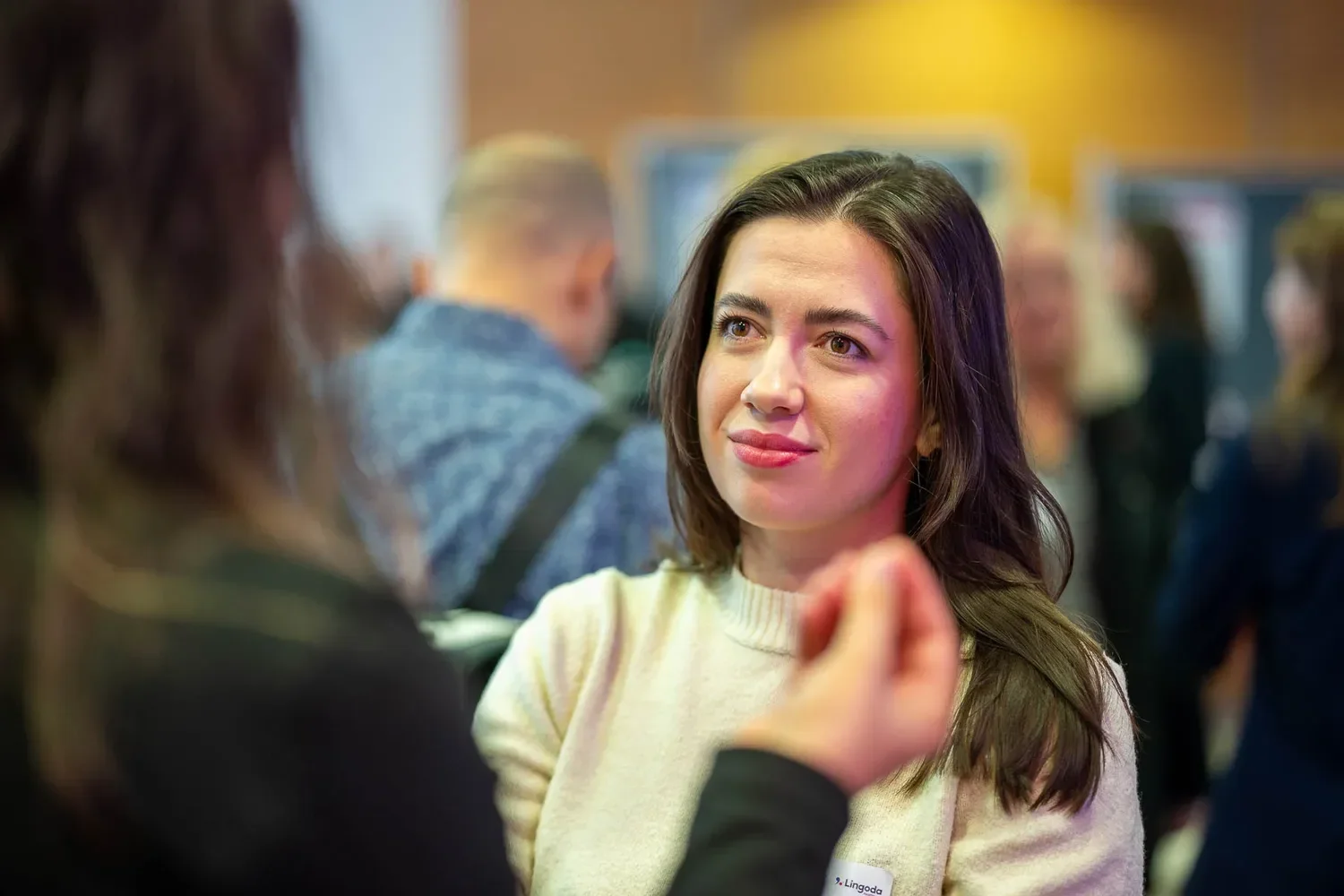 Eine junge Frau mit braunen Haaren und einem weißen Pullover schaut nachdenklich während eines Gesprächs auf einer Veranstaltung. Im Hintergrund sind weitere Menschen, die an einem Messe- oder Konferenz-Event teilnehmen.