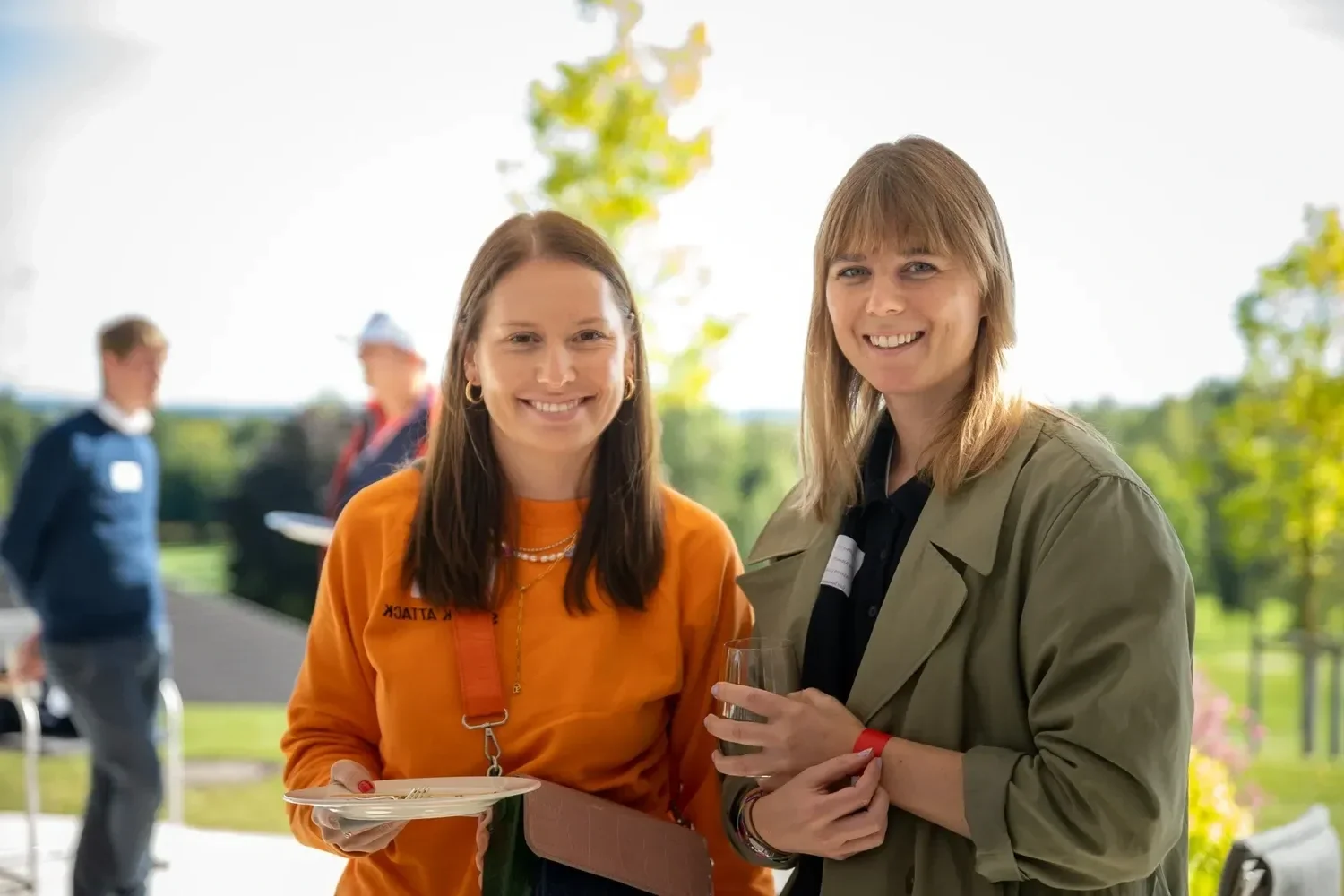 Zwei Frauen, die lächelnd im Freien bei einer gesellschaftlichen Veranstaltung stehen, eine mit einem Glas Wein, die andere mit einem Teller und Notizblock, im Hintergrund sind zwei weitere Personen.