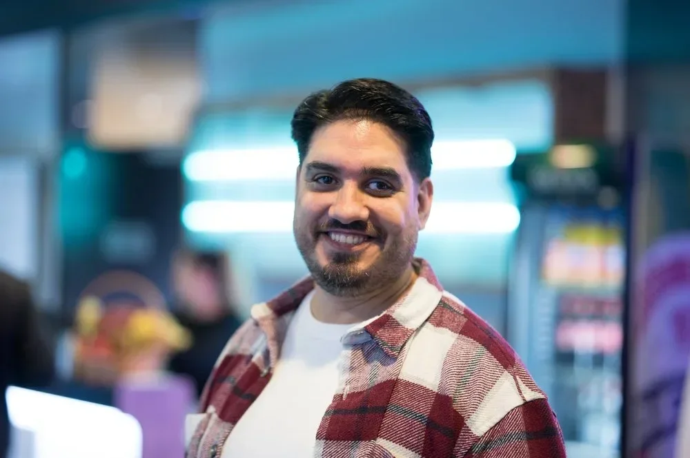 Ein Mann mit dunklem Haar, Bart und einem Lächeln, trägt ein kariertes Hemd, im Hintergrund eine helle Neonbeleuchtung und eine Cafékette.