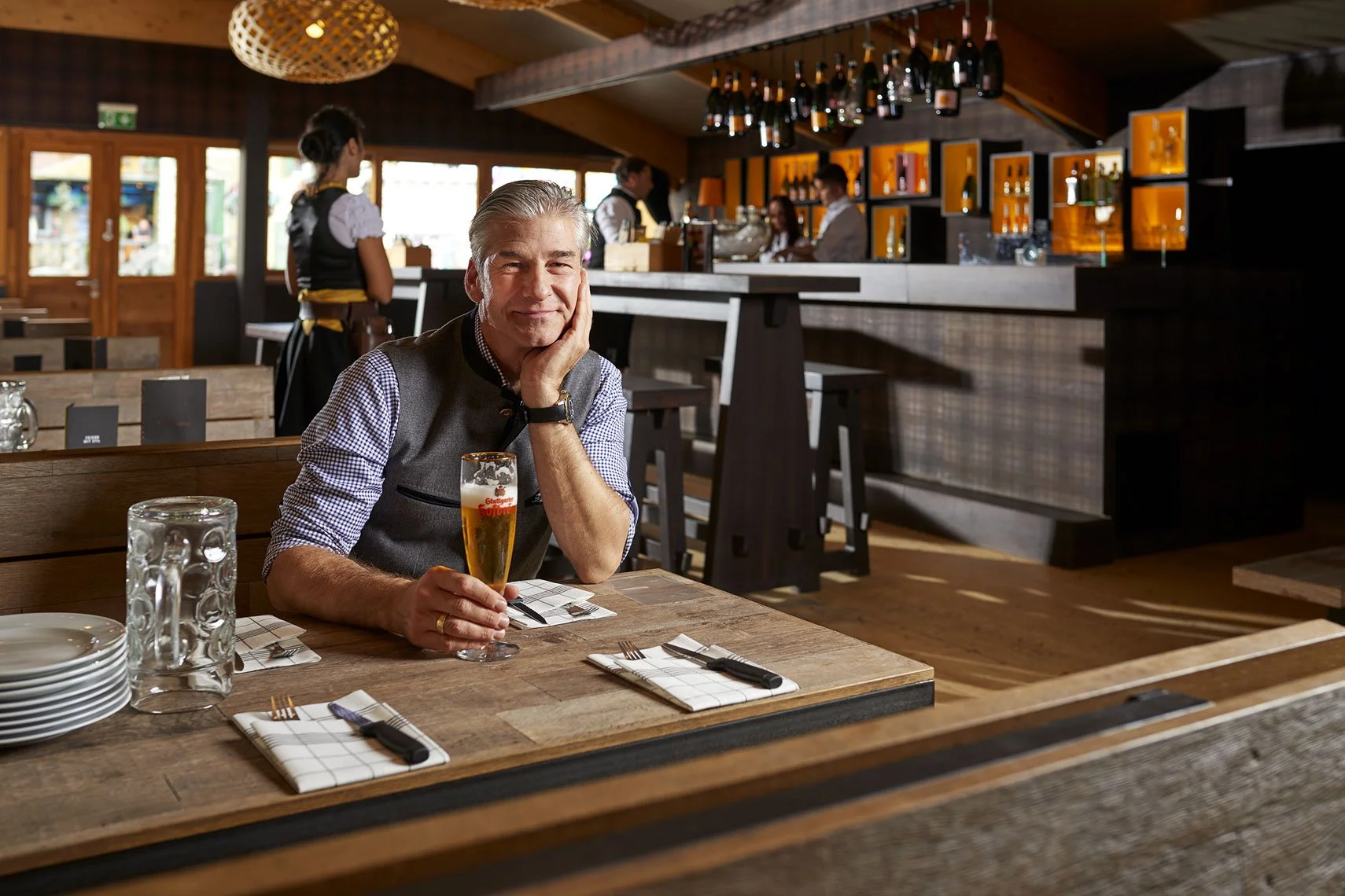 Ein älterer Mann sitzt in einem gemütlichen Restaurant mit Holzdeko, hält ein Glas Bier in der Hand, während im Hintergrund Kellner und Barpersonal zu sehen sind.