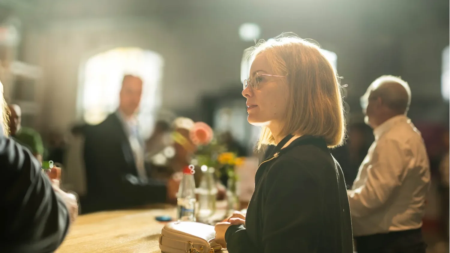 Frau mit Brille und Laptops sitzt in einem hell erleuchteten Raum, umgeben von anderen Personen, bei einem Meeting oder einer Veranstaltung.