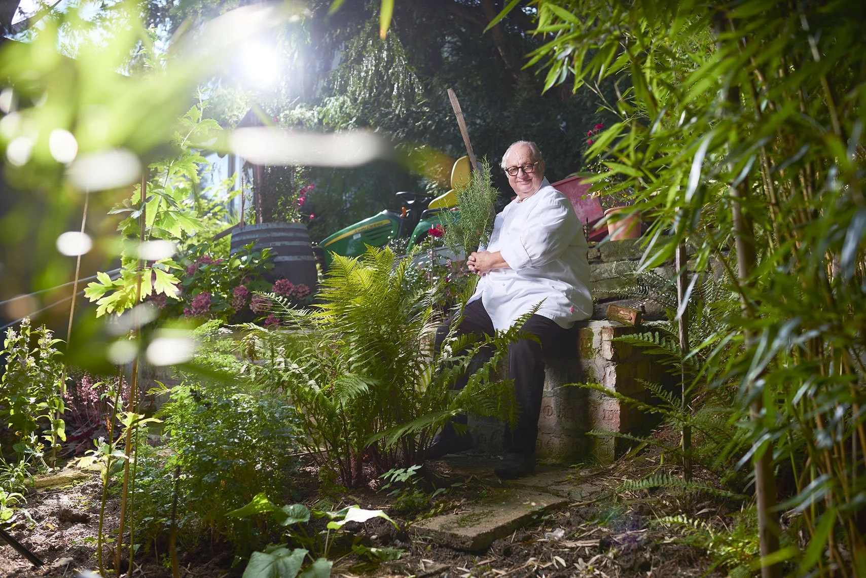 Ein Mann in weißem Kochjacke sitzt in einem grünen Garten, umgeben von Pflanzen und Blumen, hält ein Kräuterbündel in den Händen und lächelt.