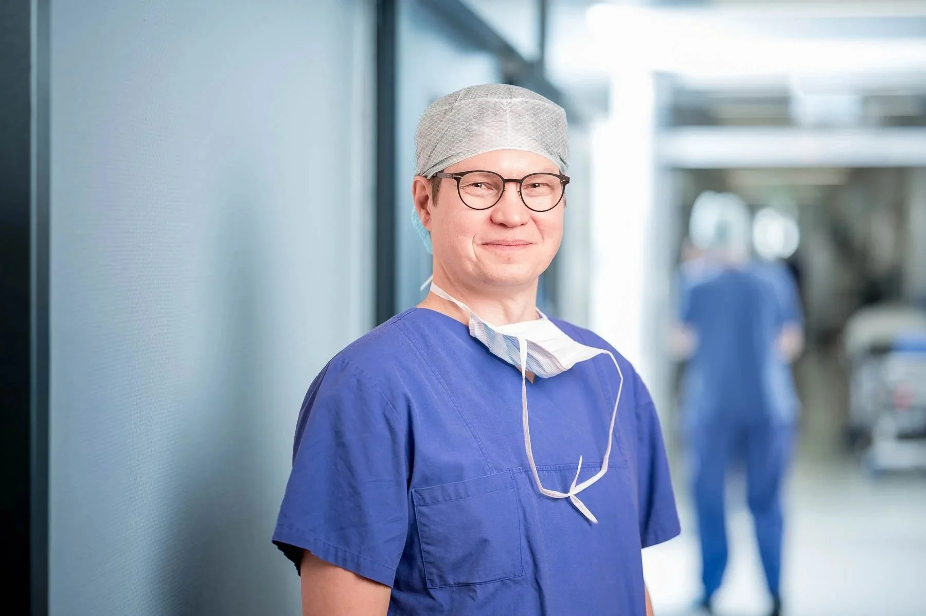 Ein männlicher Arzt oder Medizinischer Fachkraft trägt eine blaue medizinische Uniform, eine Schutzmütze und eine Maske, die um den Hals hängt, in einem Krankenhausflur.