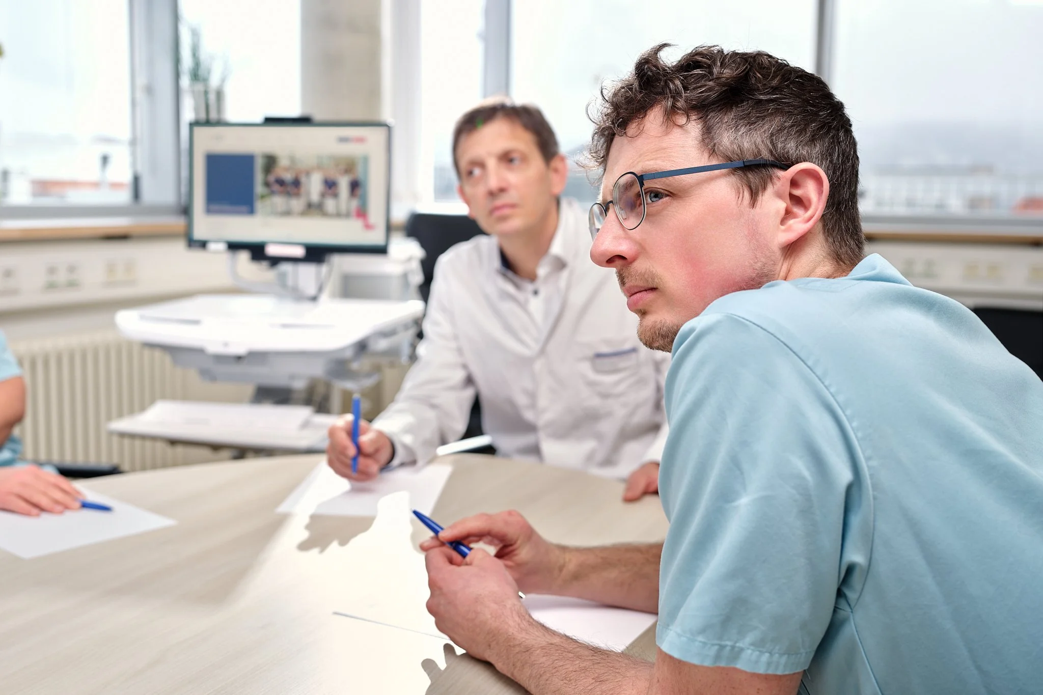 Zwei Männer in einem Büro bei einer Besprechung, einer trägt eine blaue medizinische Kluft und trägt eine Brille, der andere trägt ein weißes Hemd. Sie sitzen an einem Tisch und schauen aufmerksam.