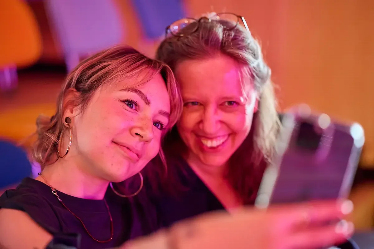 Zwei lachende Frauen machen ein Selfie in einem Raum mit warmem Licht.