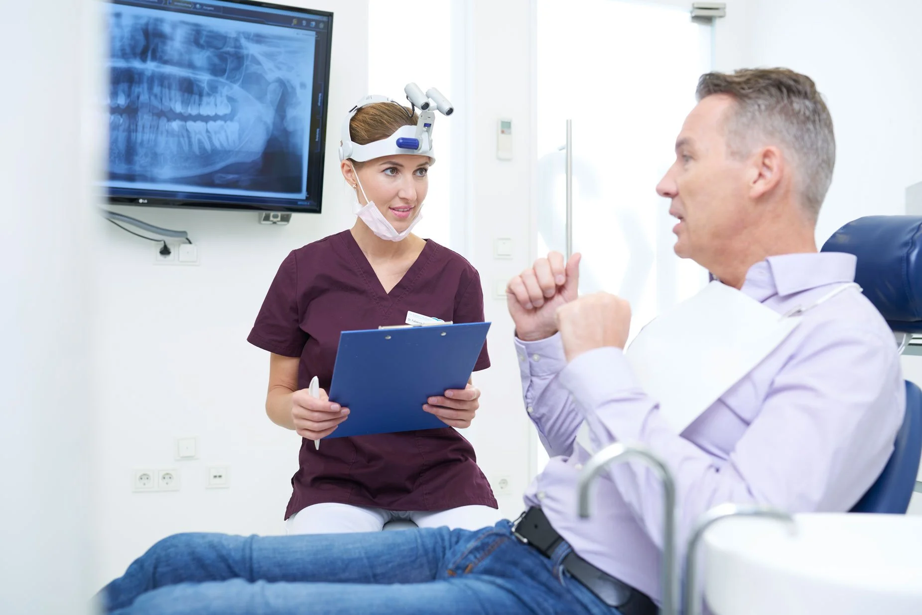 Ein Arzt spricht mit einem Patienten im Behandlungszimmer, während eine Krankenschwester das Röntgenbild eines Schädels zeigt.