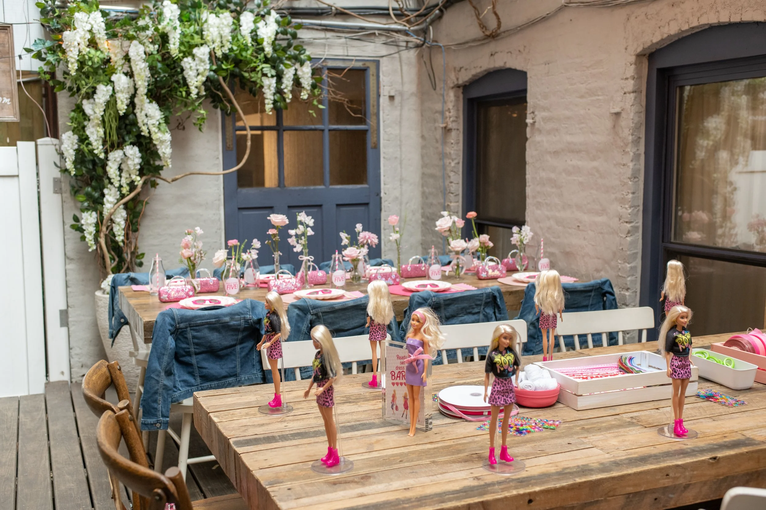 A birthday party setup for a young girl featuring a table decorated with pink and white party supplies, Barbie dolls, and a backdrop with flowers and a blue door.