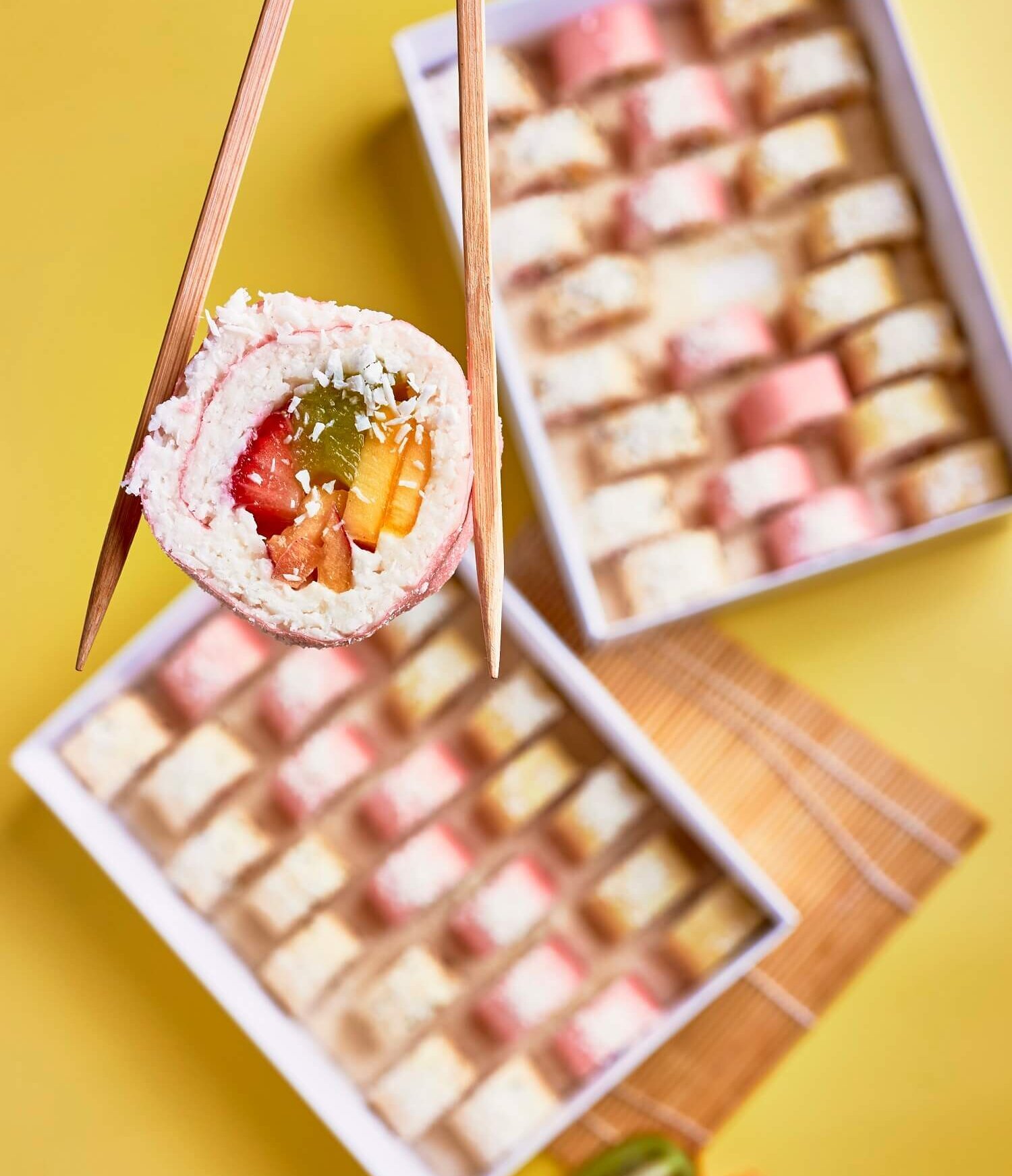 A piece of fruit-filled Swiss roll cake held with chopsticks over a yellow background, with two open boxes of assorted miniature cakes or pastries in the background.