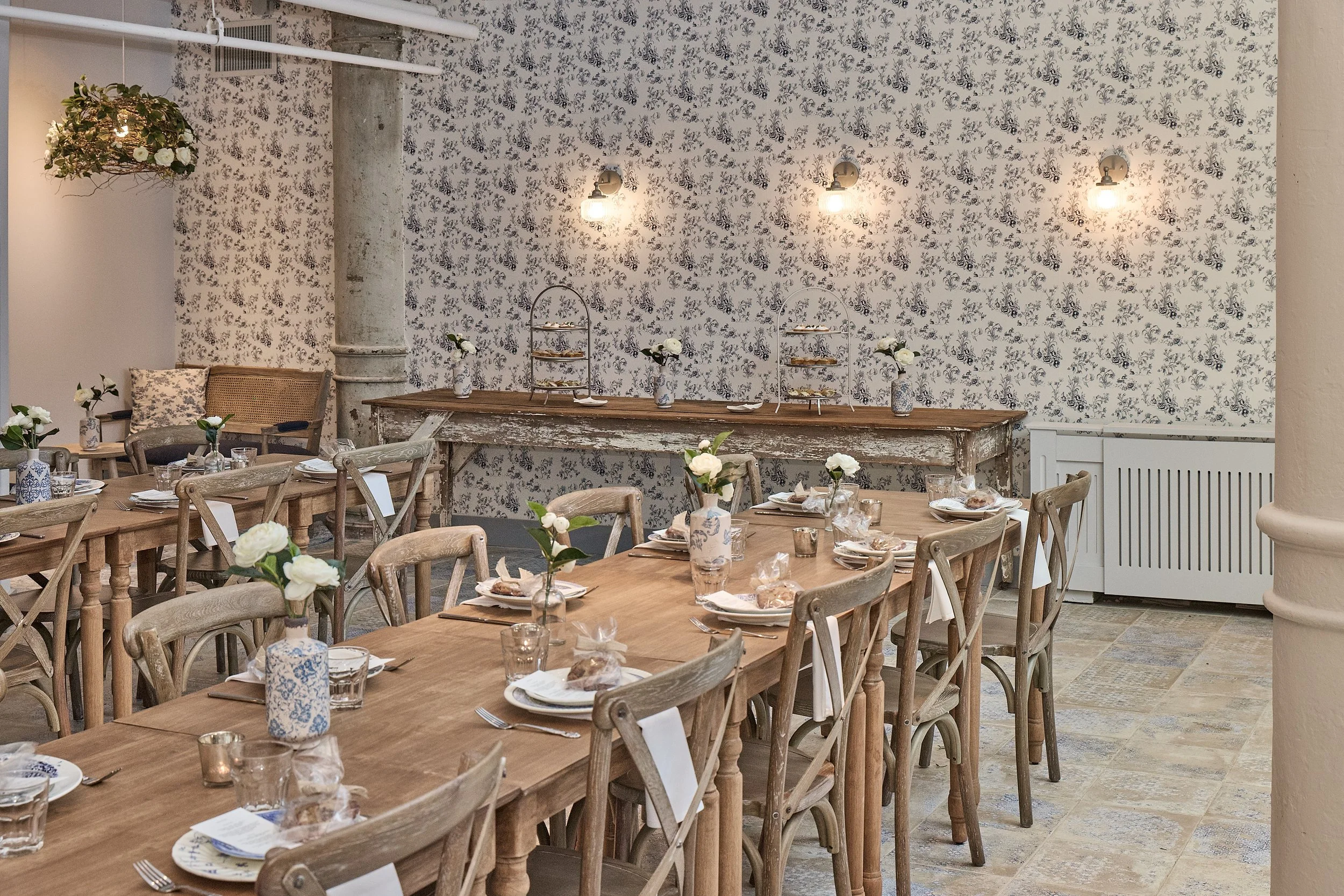 A dining room with a rustic wooden table set with plates, glasses, and wrapped bread rolls, surrounded by wooden chairs, decorated with white flowers in blue and white vases, against a wall with black and white floral wallpaper.