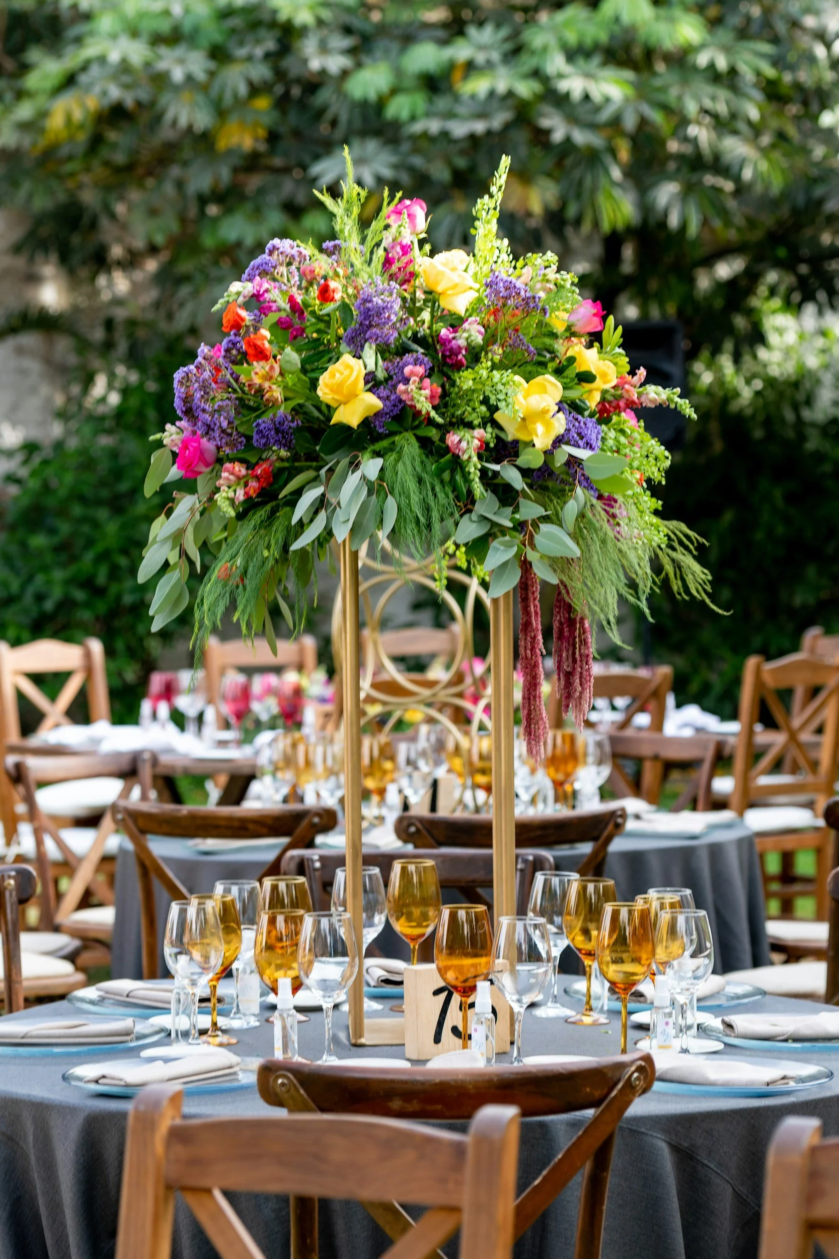 Elegant outdoor dining table with a large colorful flower arrangement as the centerpiece, set with gold-tinted glasses, white napkins, and neatly arranged plates, surrounded by wooden chairs in a garden setting.