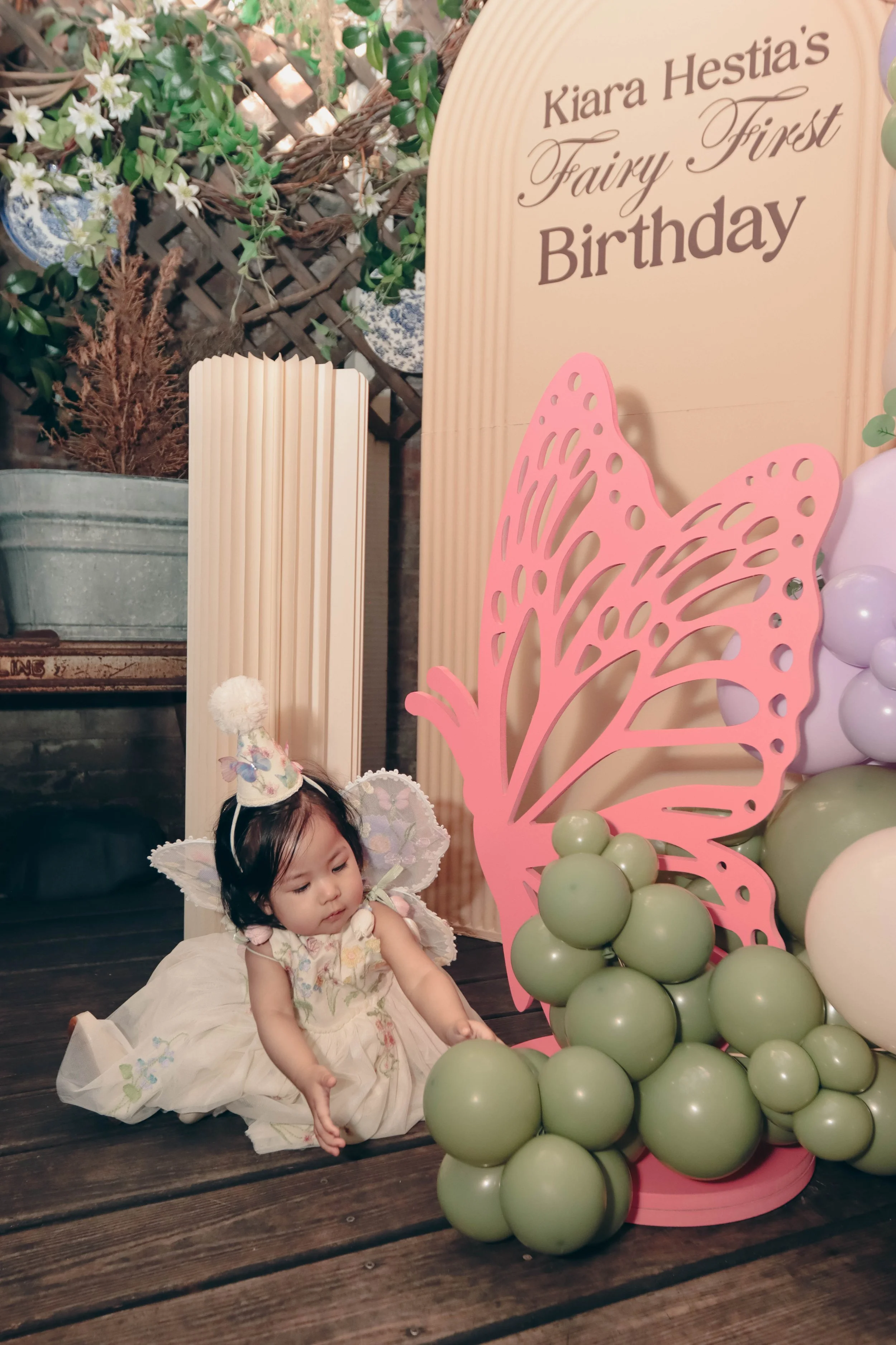 A young girl dressed as a fairy with wings and a floral dress, sitting on a wooden floor next to a pink butterfly-shaped decoration with green balloons, celebrating Kiara Hestia's fairy-themed first birthday.