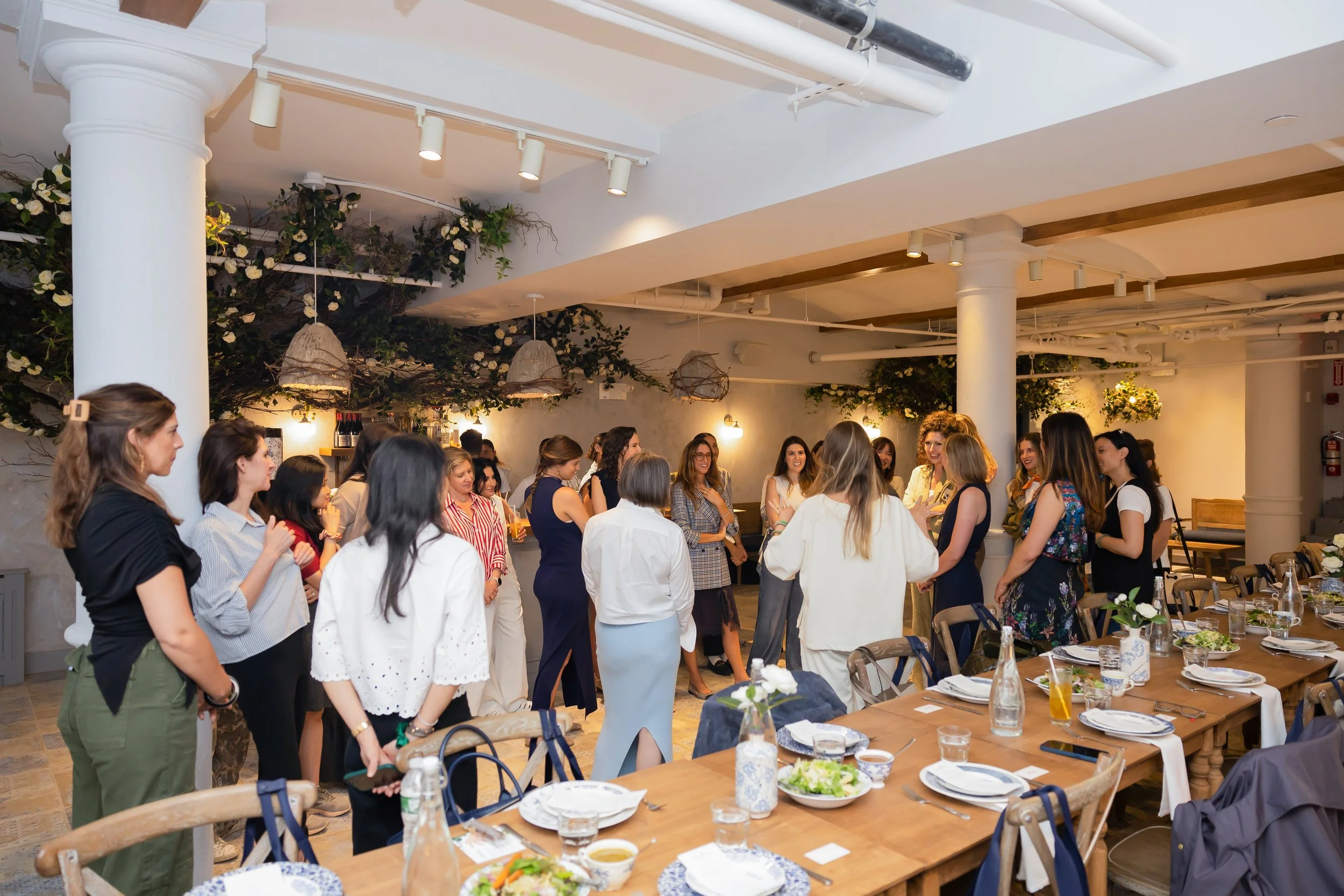 Group of women gathered in a restaurant dining area, socializing and talking, with tables set for a meal.