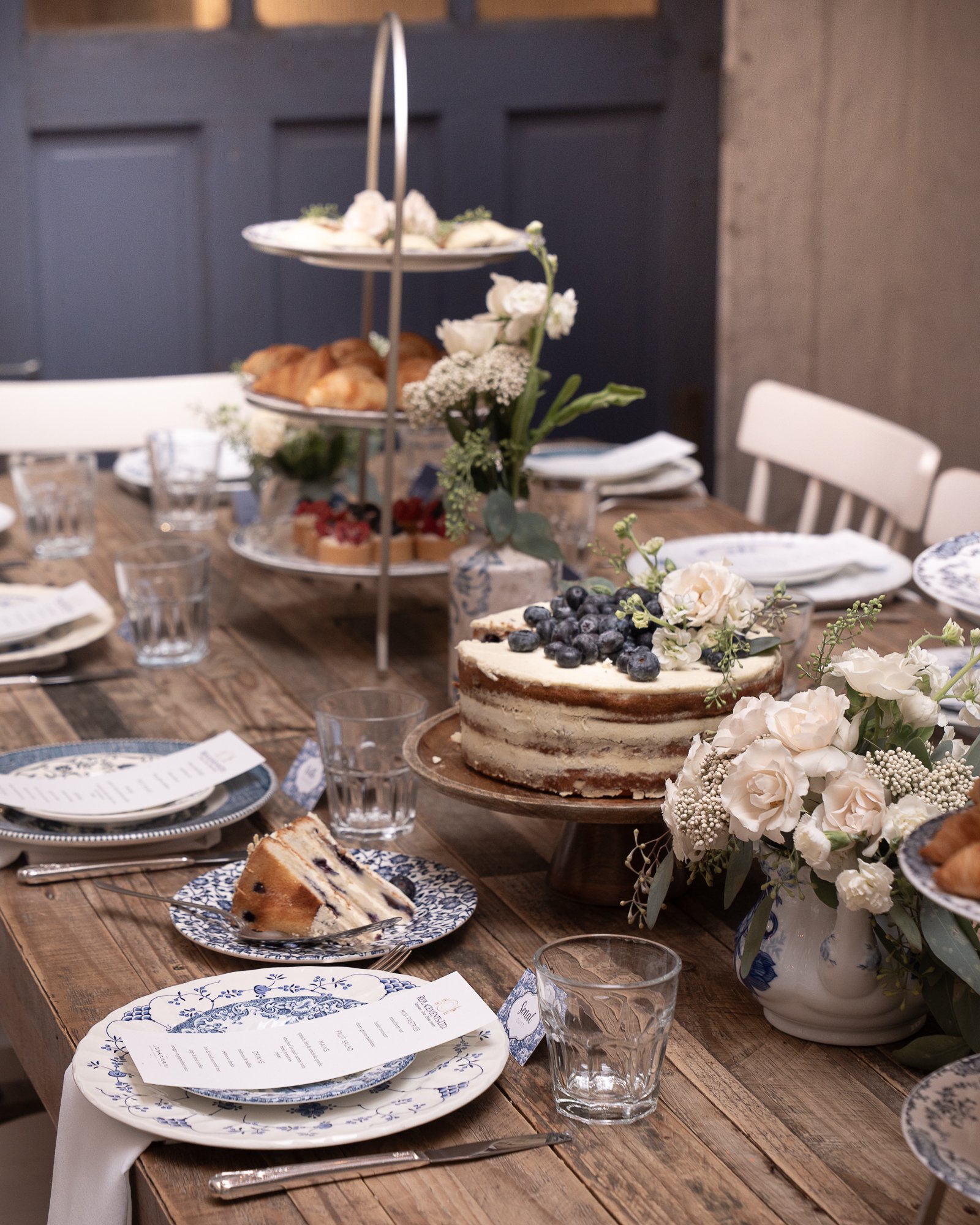 A rustic dining table set with floral arrangements, a layered cake topped with blueberries, plates, menus, and glasses, with a three-tiered serving stand in the background holding croissants, cupcakes, and desserts.