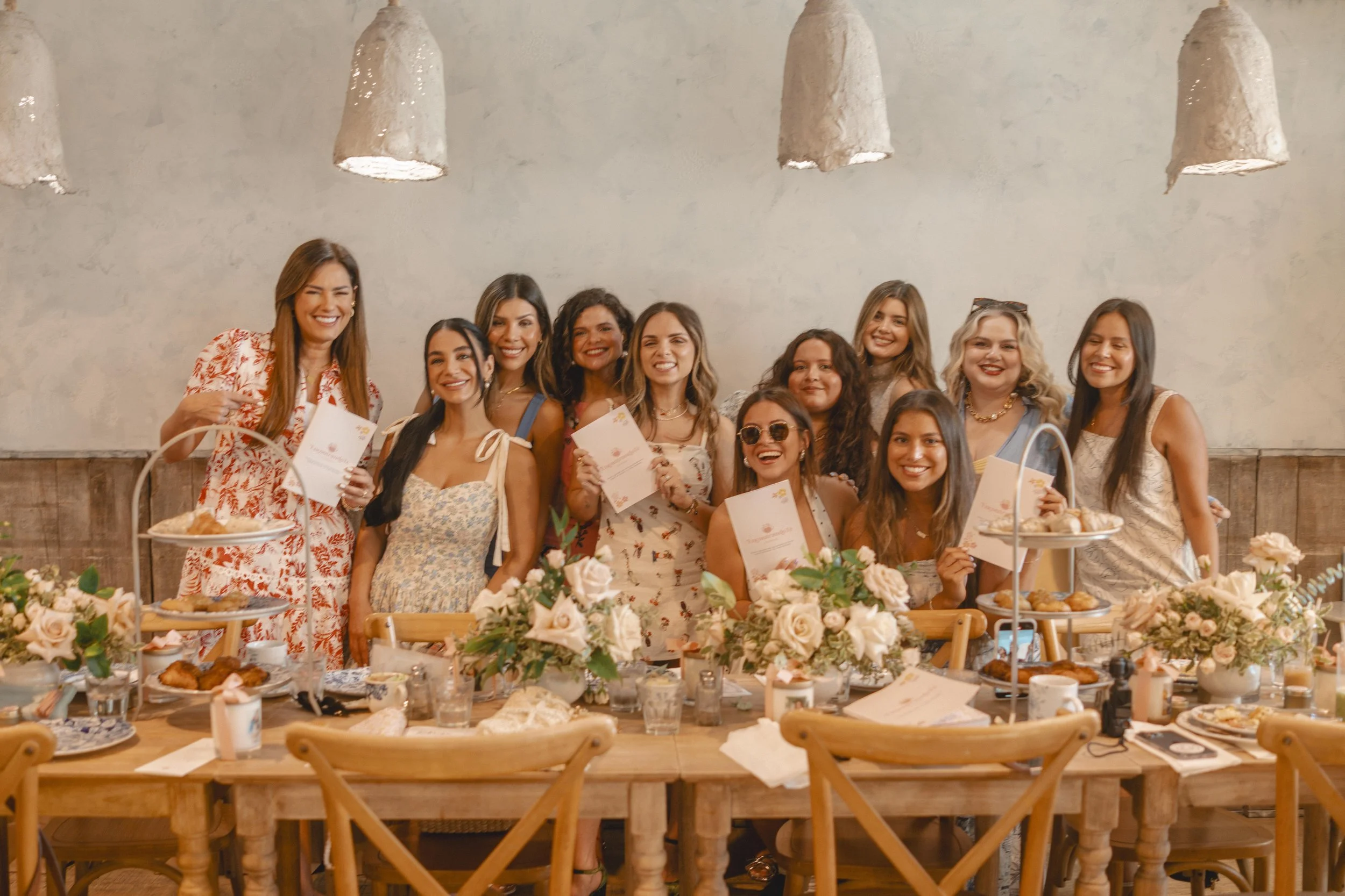 Group of women celebrating at a wedding or party, standing behind a table with floral arrangements and desserts, holding certificates and smiling for the camera.