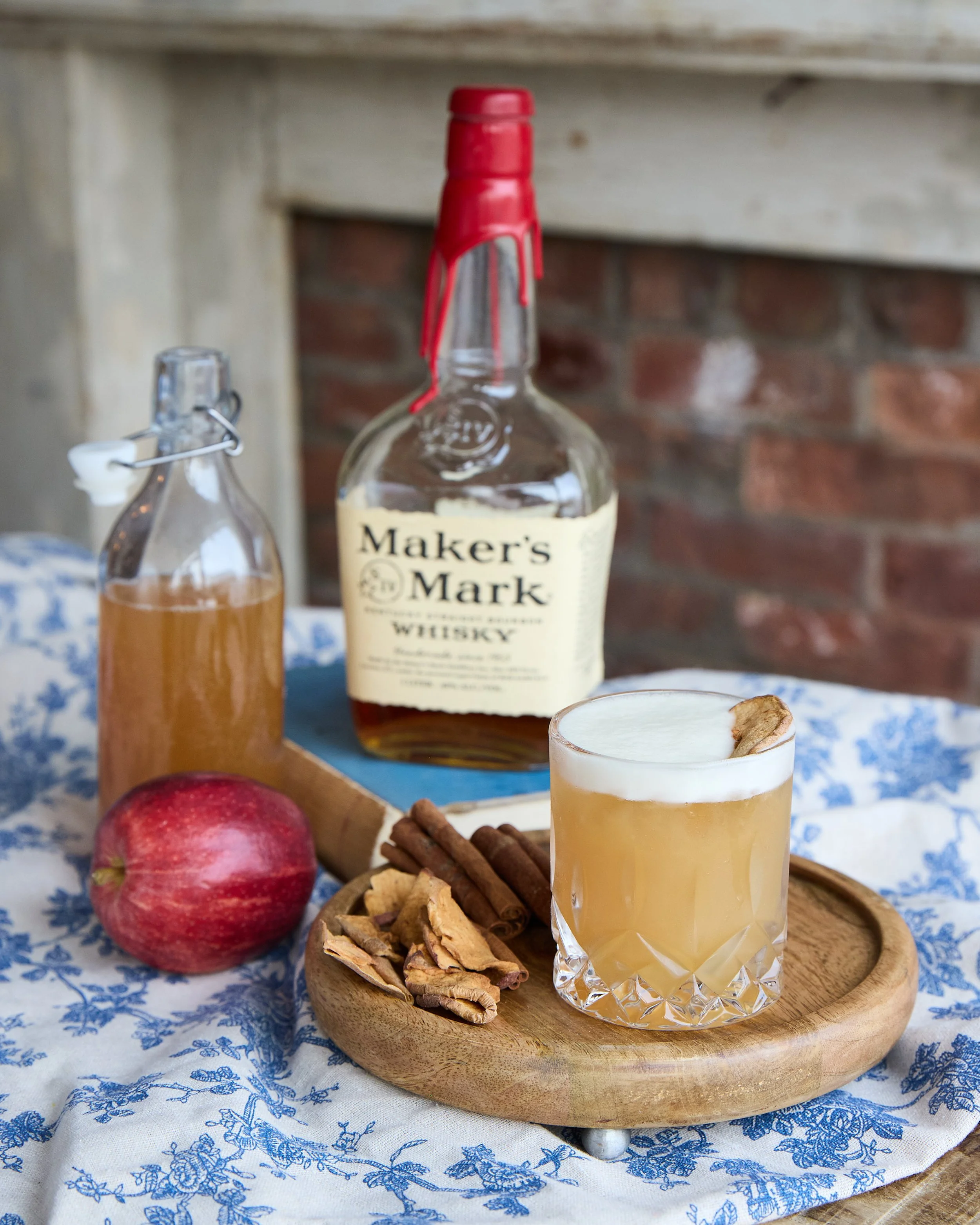 A glass of whiskey cocktail garnished with a ginger slice, placed on a wooden tray with cinnamon sticks, ginger, apple, and a bottle of Maker's Mark whiskey in the background.